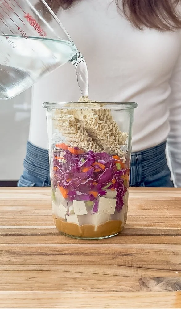 Layered glass jar with ingredients for instant noodles and boiling water being poured over the top.