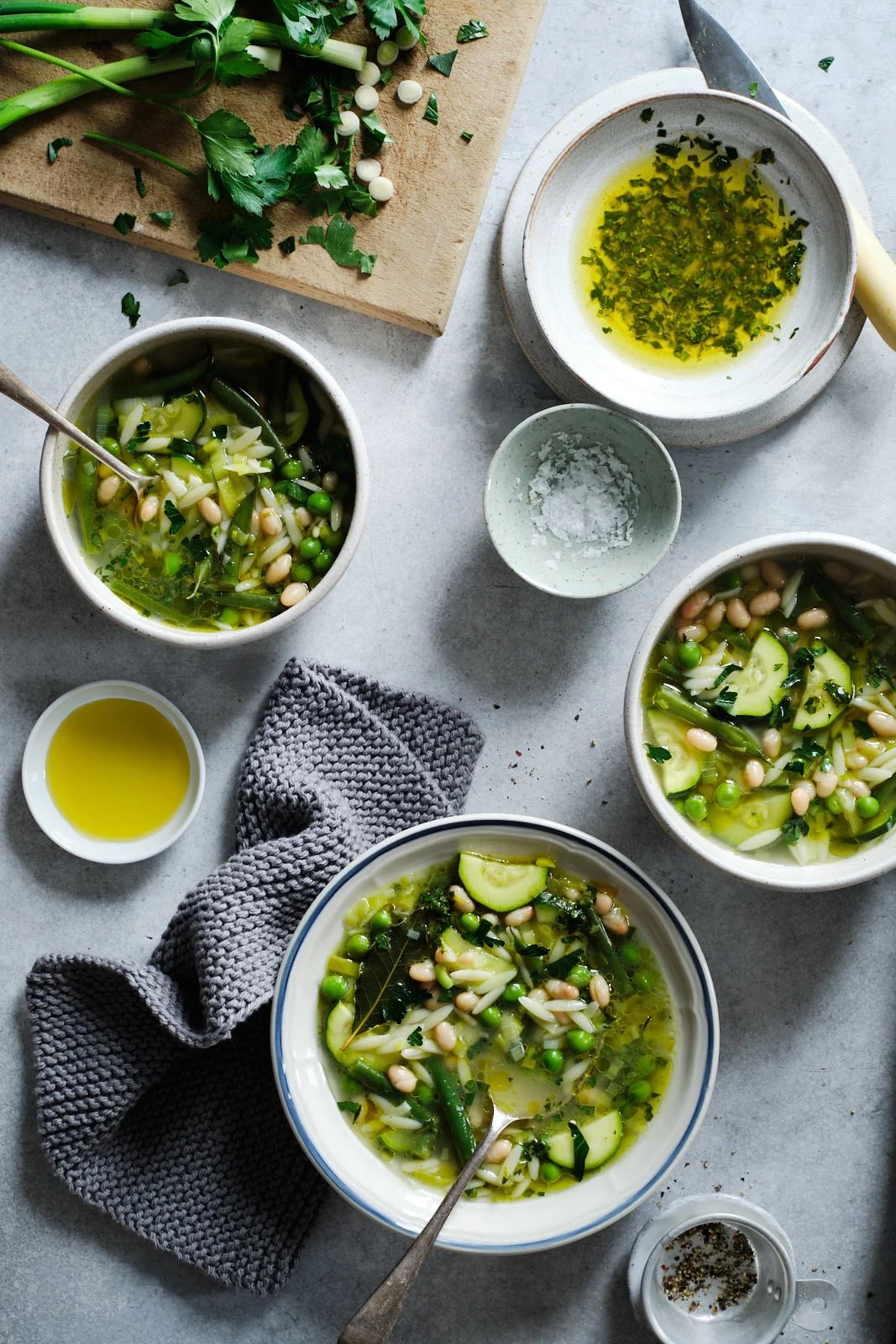 Three bowls with green minestrone soup, a vegetarian soup made with zucchini, green beans, peas and white beans. Also in frame bowls with condiments and a cutting board with chopped green onions.