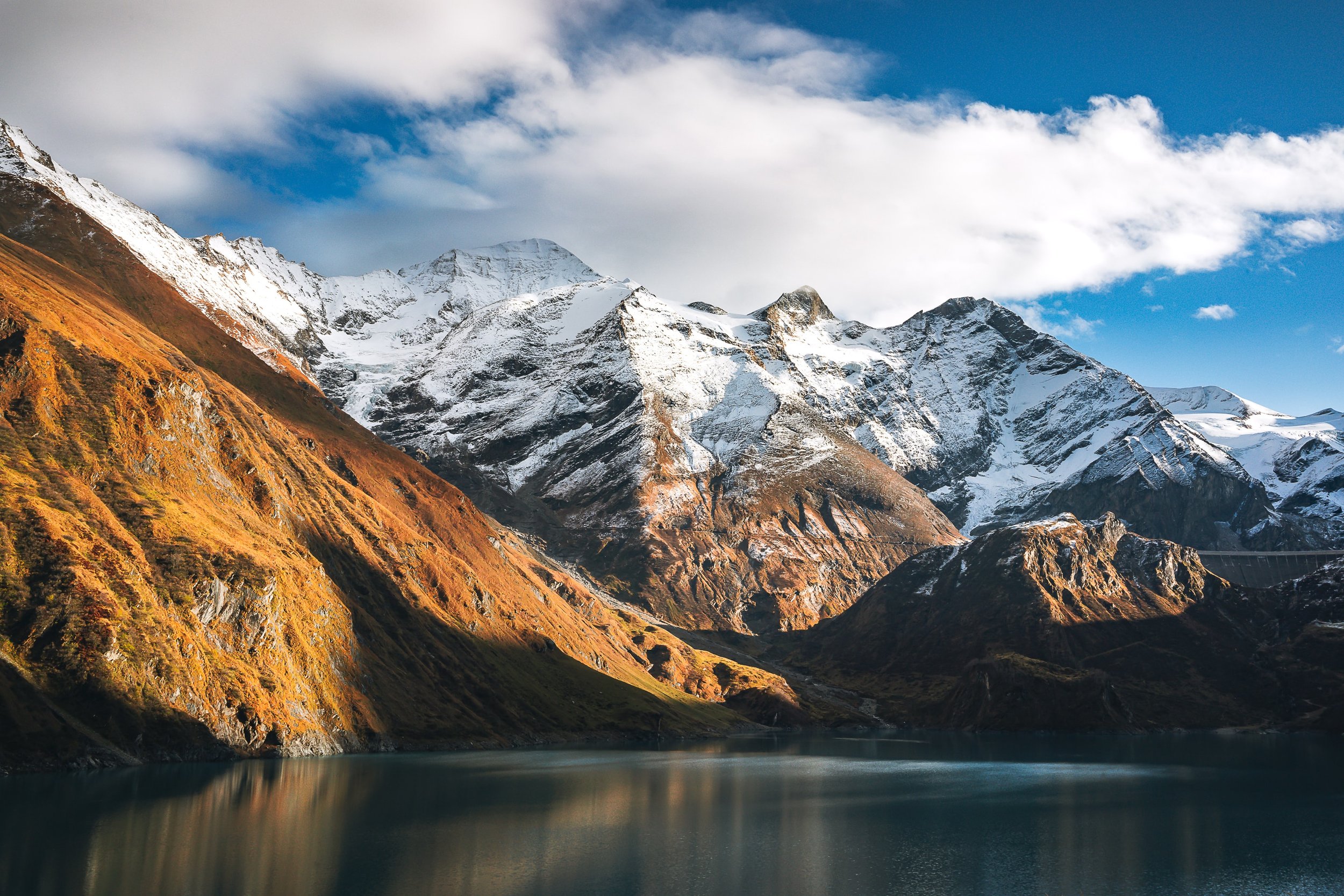 Image of a mountain lake with calm waters and a snowcapped mountain range with puffy white clouds and a blue sky.