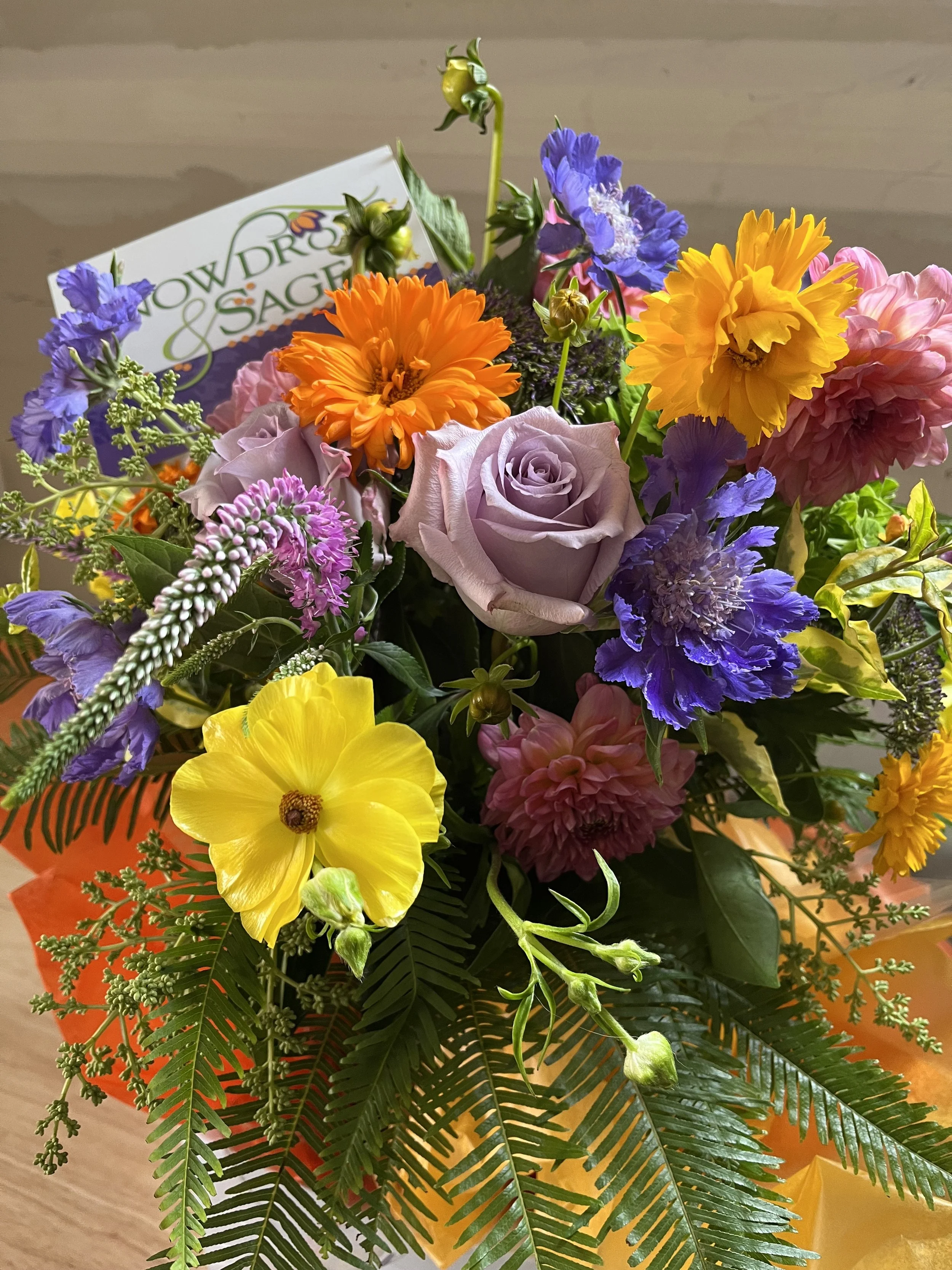 Bright, cheerful arrangement with yellow, orange, and purple flowers by Snowdrops & Sage