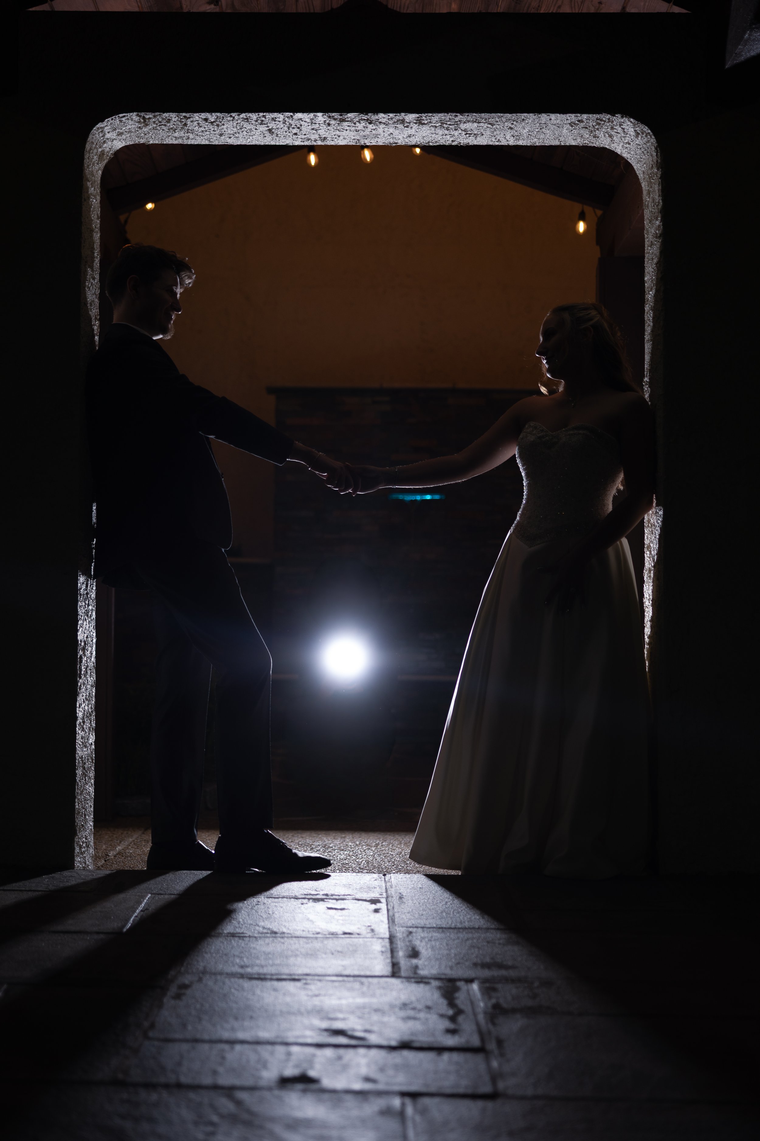 A silhouette of a couple holding hands, standing in a doorway at night. The man is dressed in a suit and the woman is wearing a strapless gown. Warm string lights are hanging in the background.