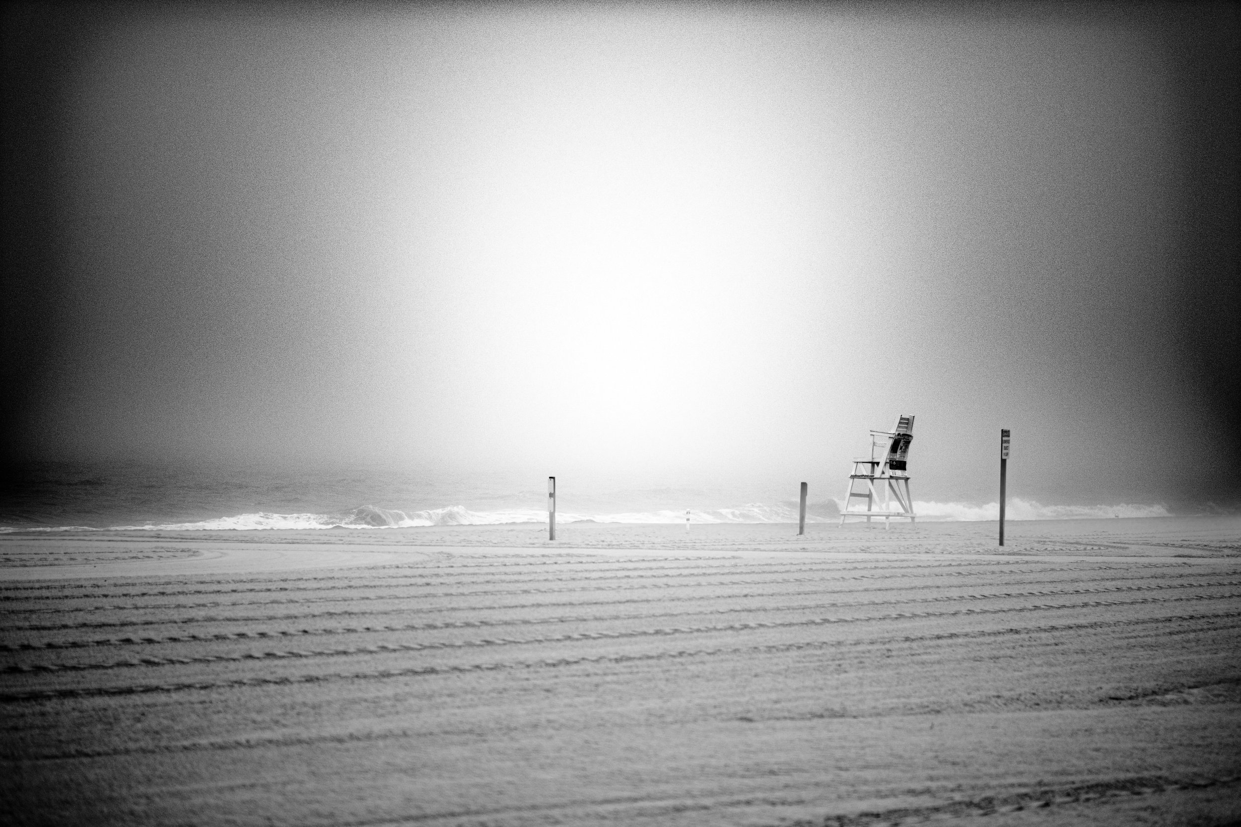 Black and white photograph of a deserted beach with a lifeguard chair and warning signs, calm ocean waves, and an overcast sky.