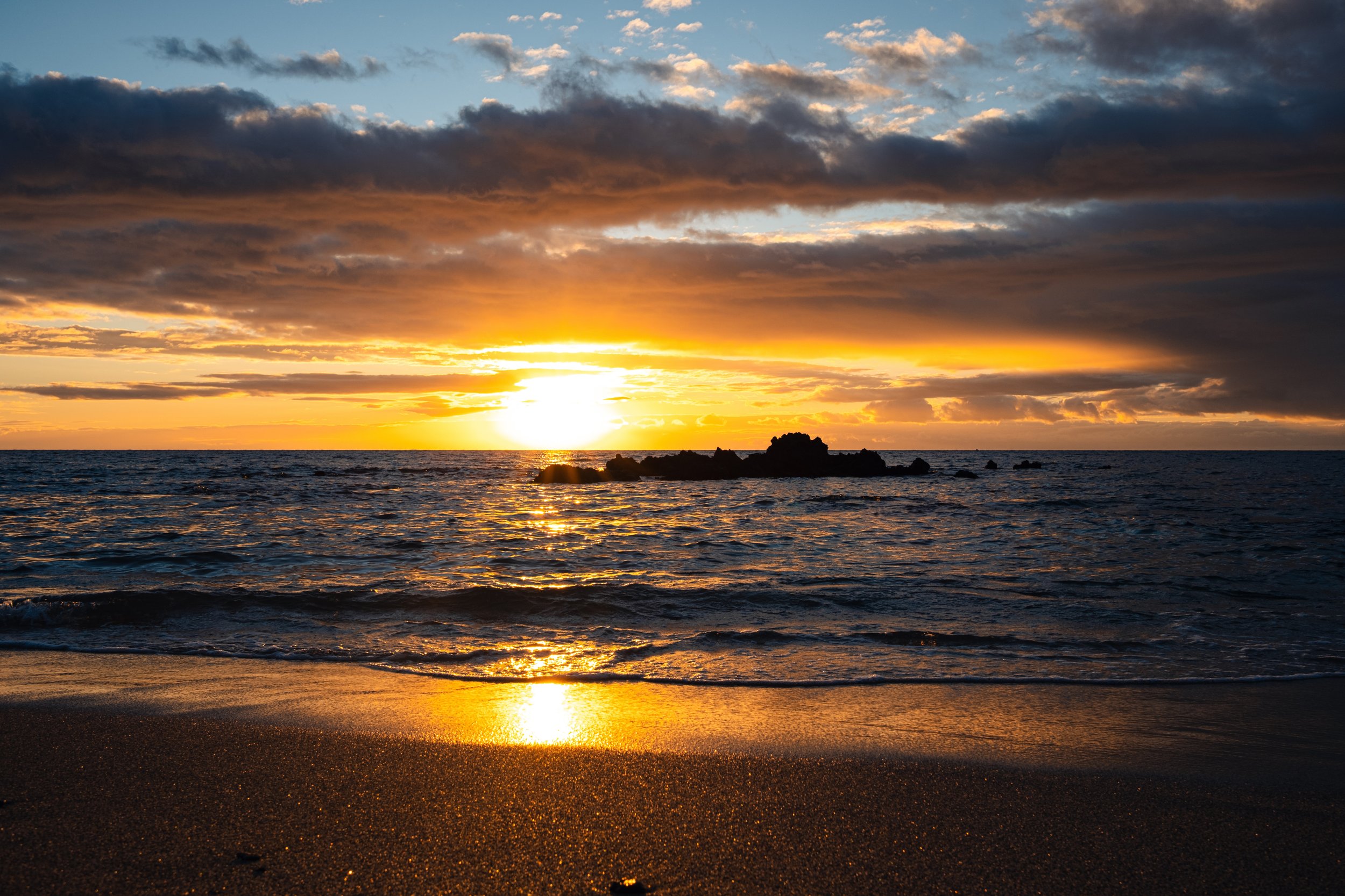 Sunset over the ocean with clouds in the sky and rocks in the water.