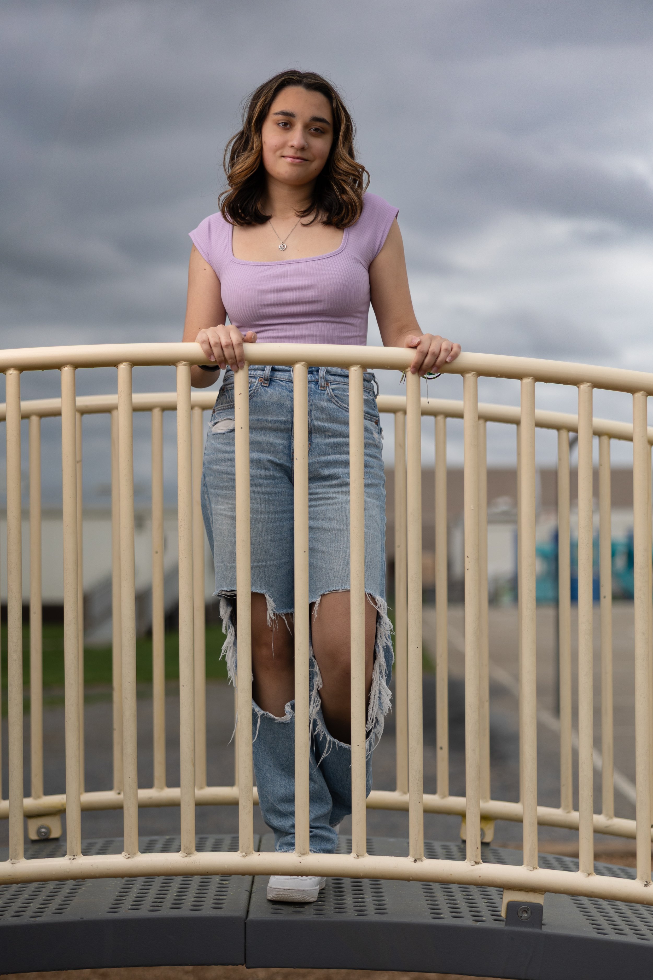Young woman with wavy brown hair wearing a lavender sleeveless top and distressed blue jeans, standing on a bridge with a cloudy sky in the background.