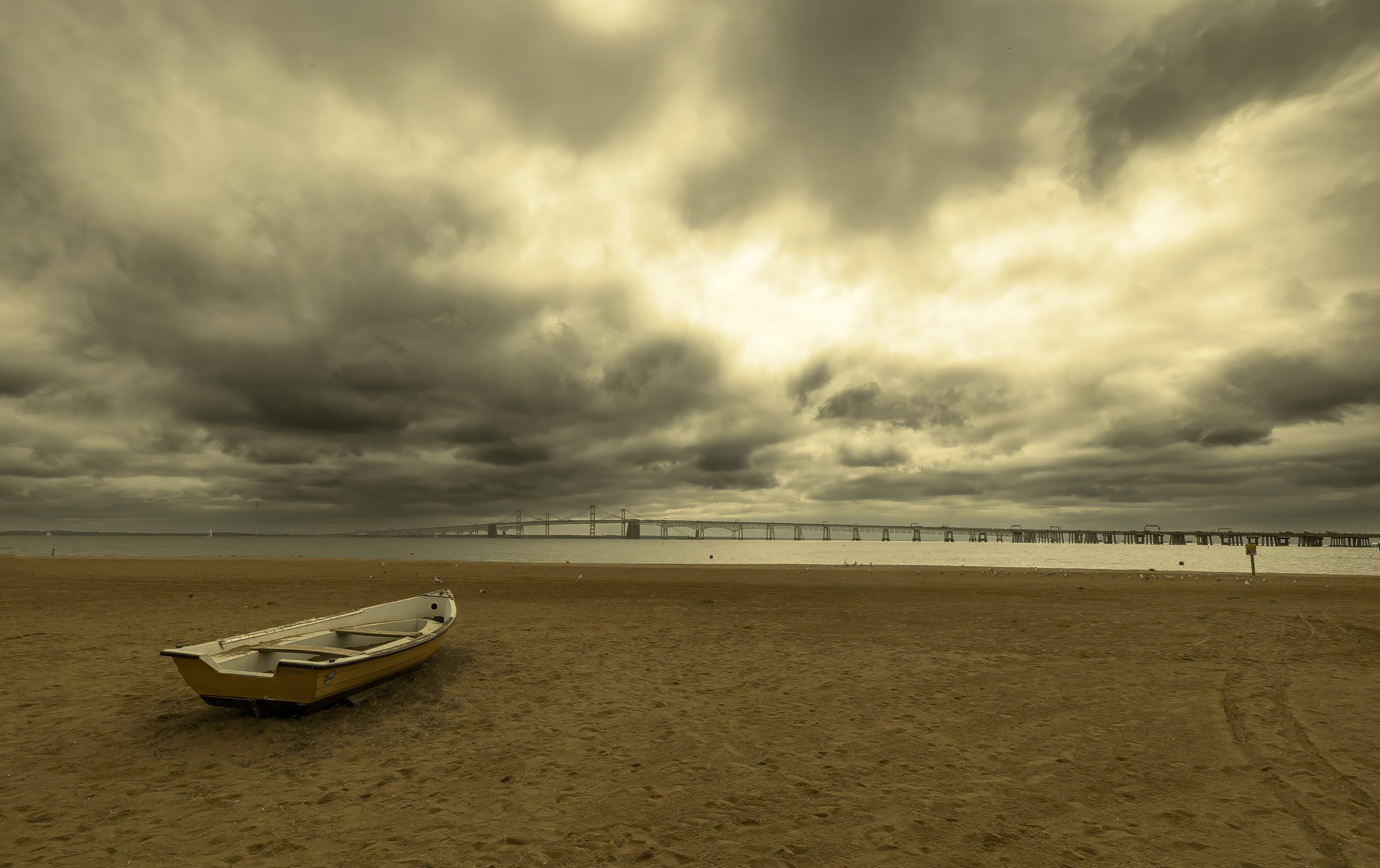 Overcast sky over a beach with a small boat on the sand and a bridge in the distance.