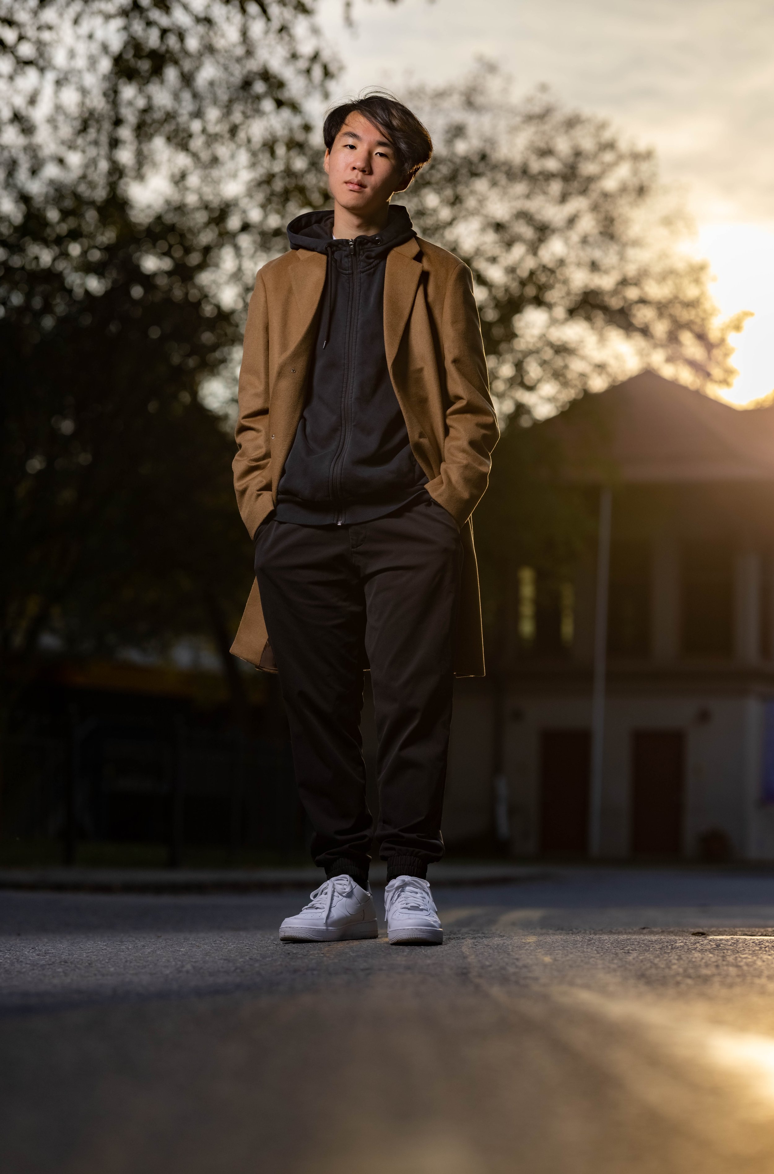 A young man walking outdoors during sunset, wearing a brown coat, black hoodie, black pants, and white sneakers, with trees and a building in the background.