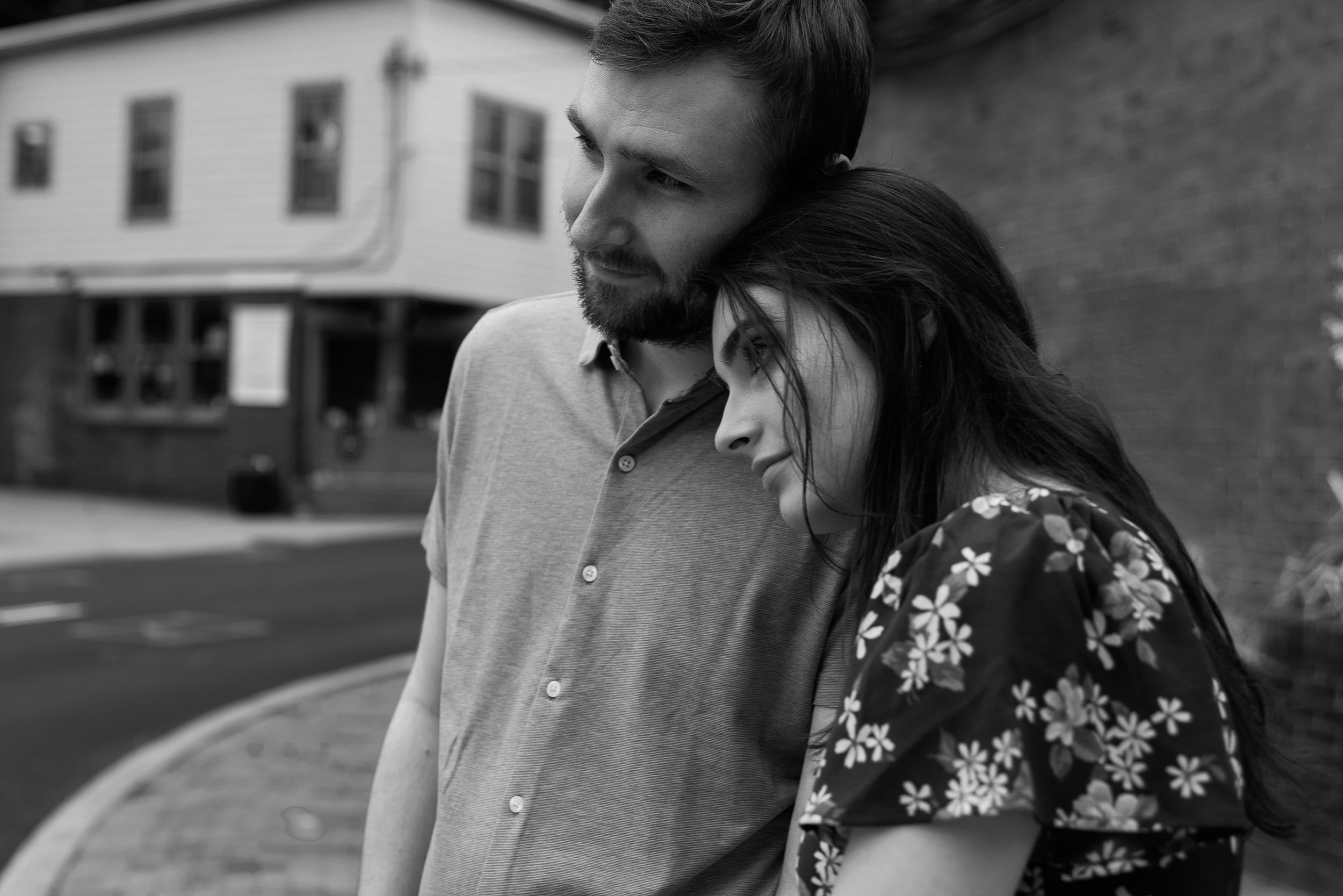 A man with a beard and a woman with long dark hair leaning their heads together outdoors in front of a house, captured in black and white.