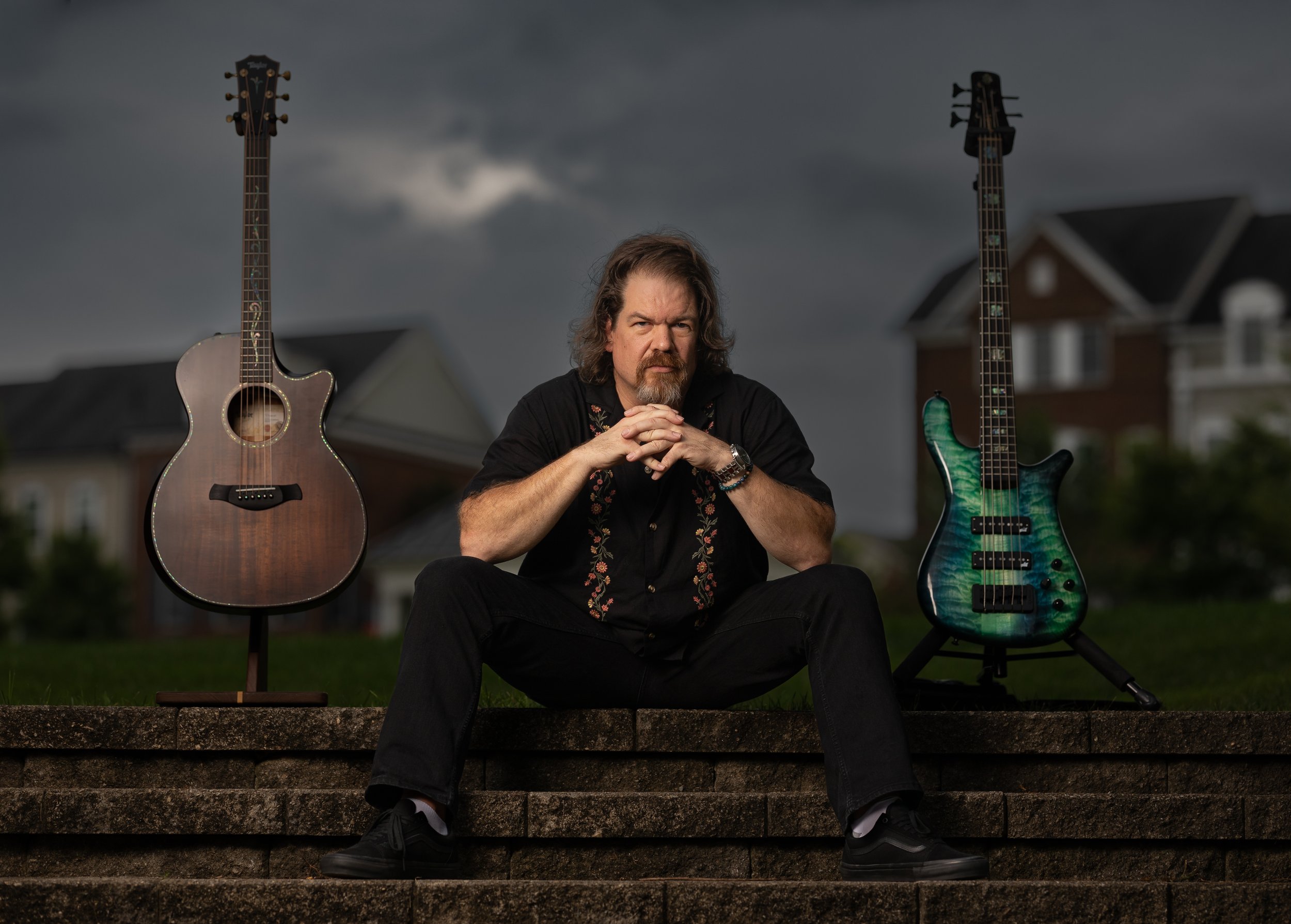 A man sitting on stone steps with two guitars, one on each side, outdoors during cloudy weather.