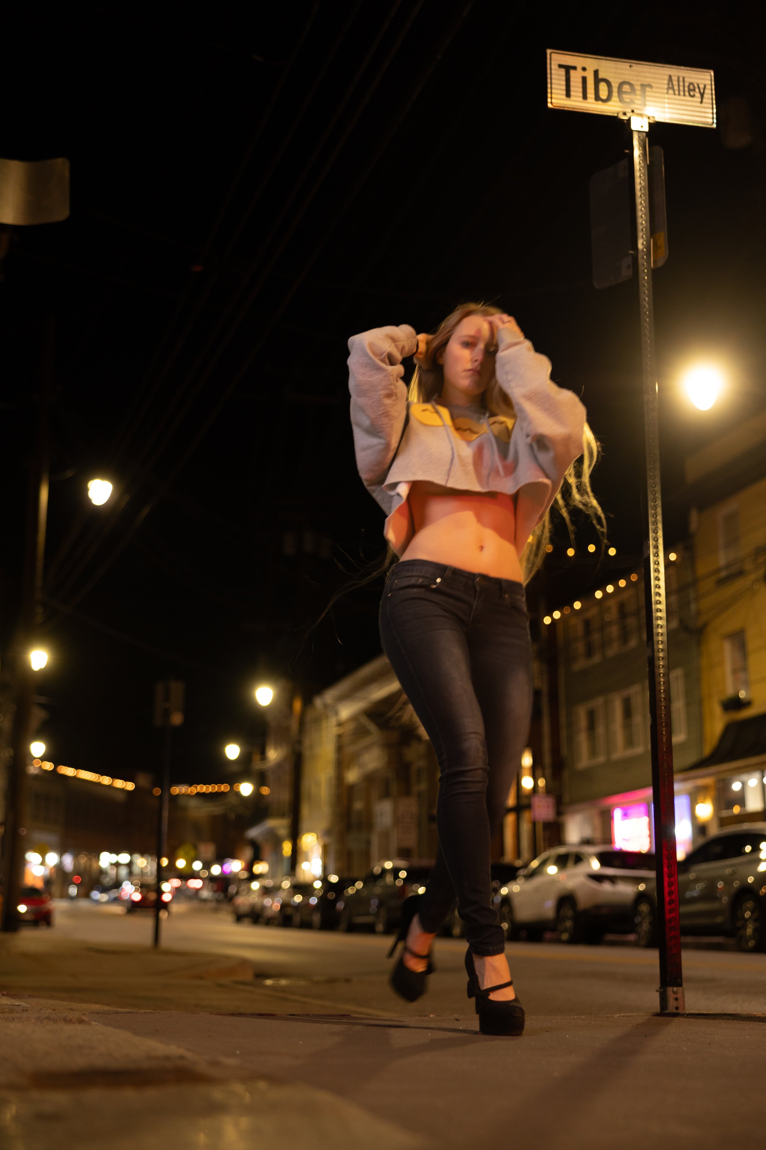 A woman walking on a city street at night, holding her hair with both hands, wearing a cropped sweatshirt and high-heeled shoes, in front of a street sign that reads 'Tiber Alley'.