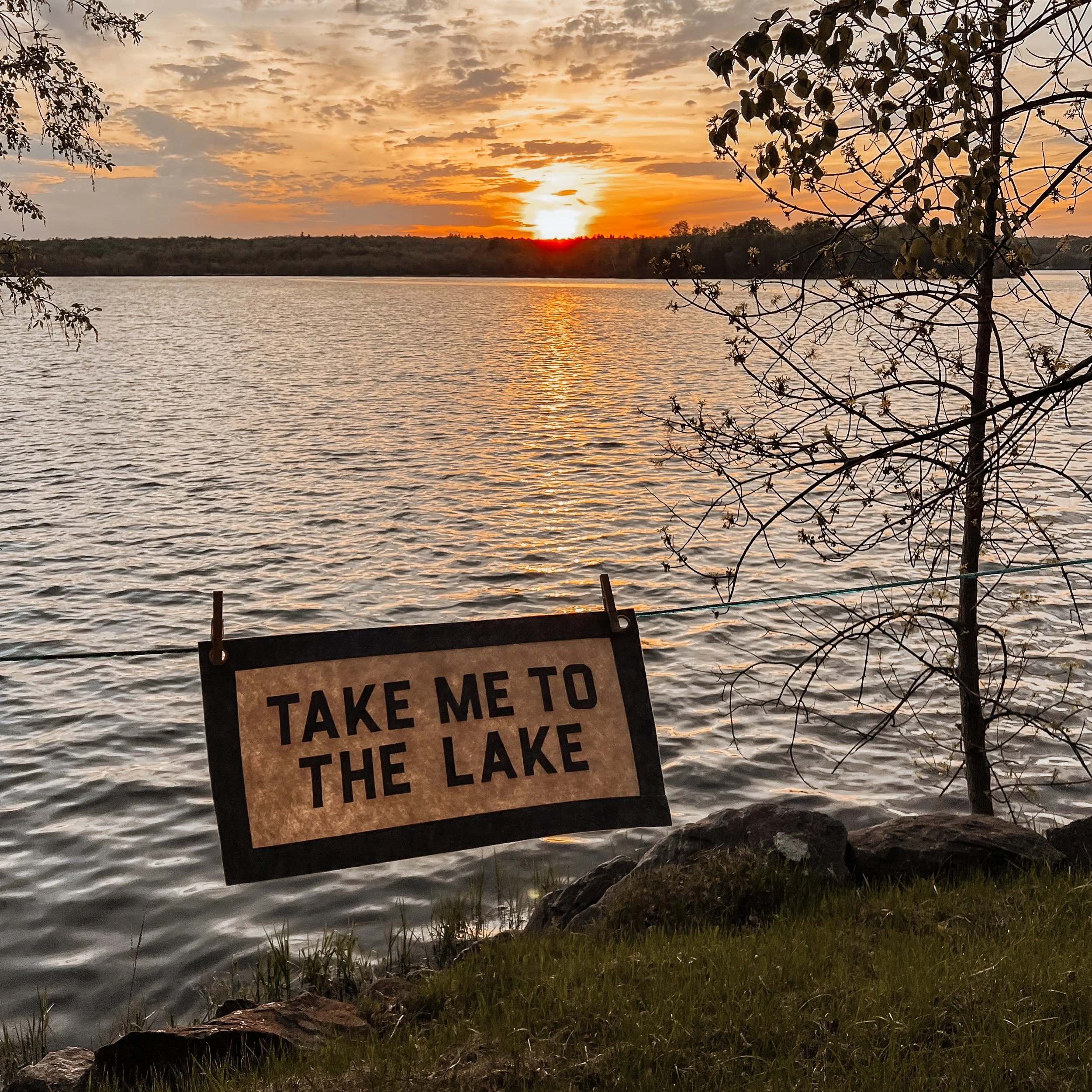 Sunset over a lake with trees on the shore and a partially visible sign in the foreground that reads 'HAVE METTA TAKE ME TO THE LAKE'.
