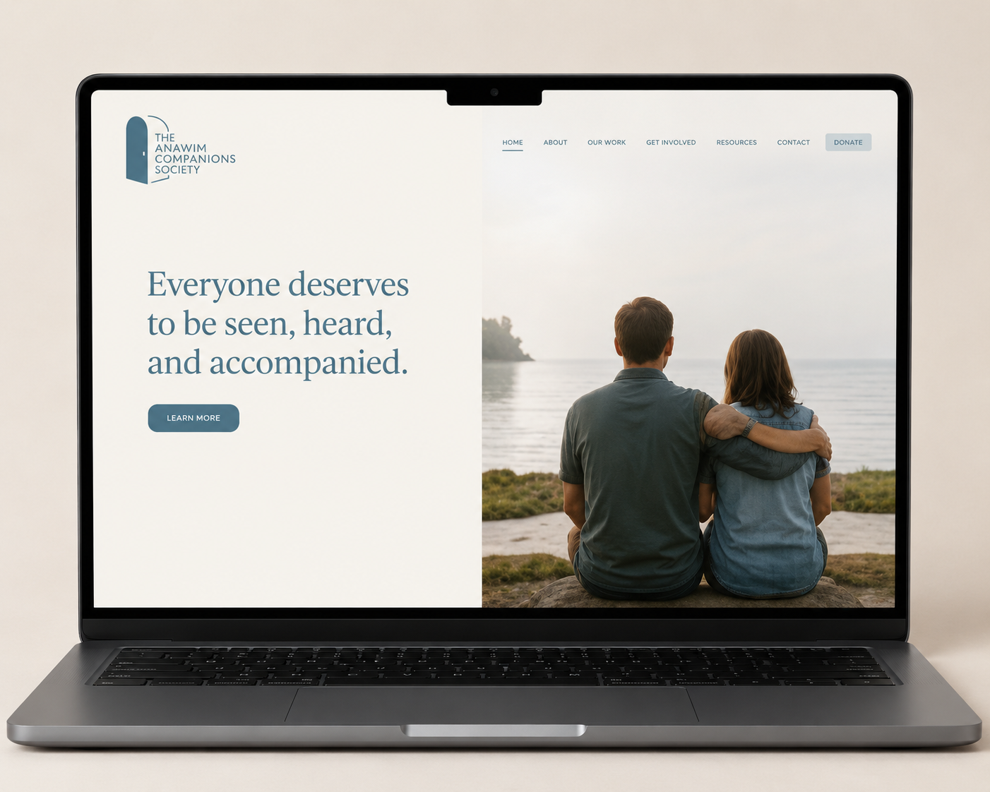 Laptop screen displaying a website for The Anawim Companions Society, showing a photo of a couple sitting by the water with their arms around each other, and the text 'Everyone deserves to be seen, heard, and accompanied.'
