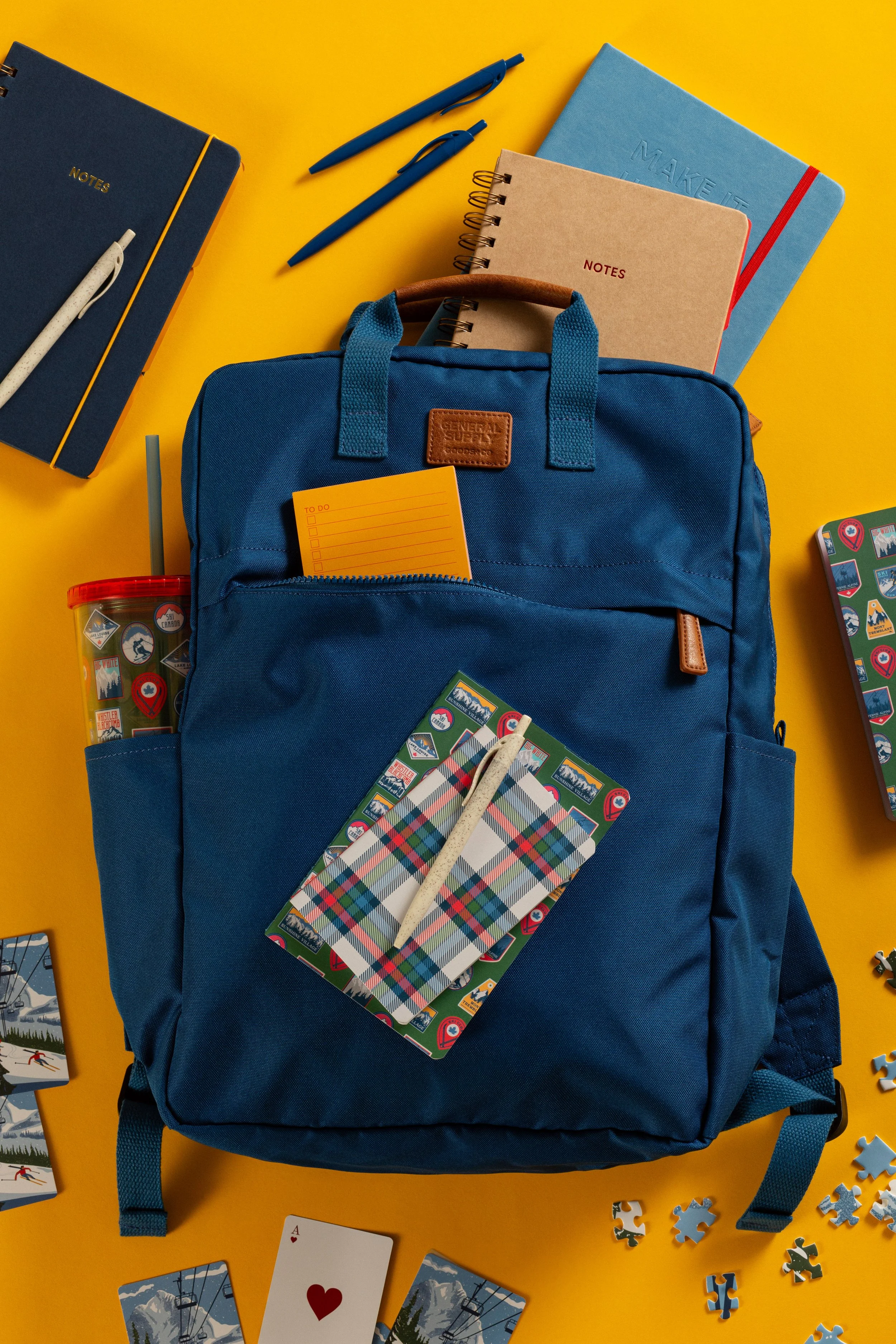 Blue backpack on a yellow surface with notebooks, pens, a container, playing cards, and scattered puzzle pieces.