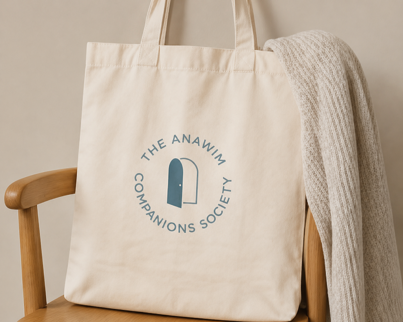A beige tote bag hanging on the back of a wooden chair with a light-colored knit blanket draped over the chair. The tote bag has a logo that reads 'The Anawin Companions Society' with an open door icon.
