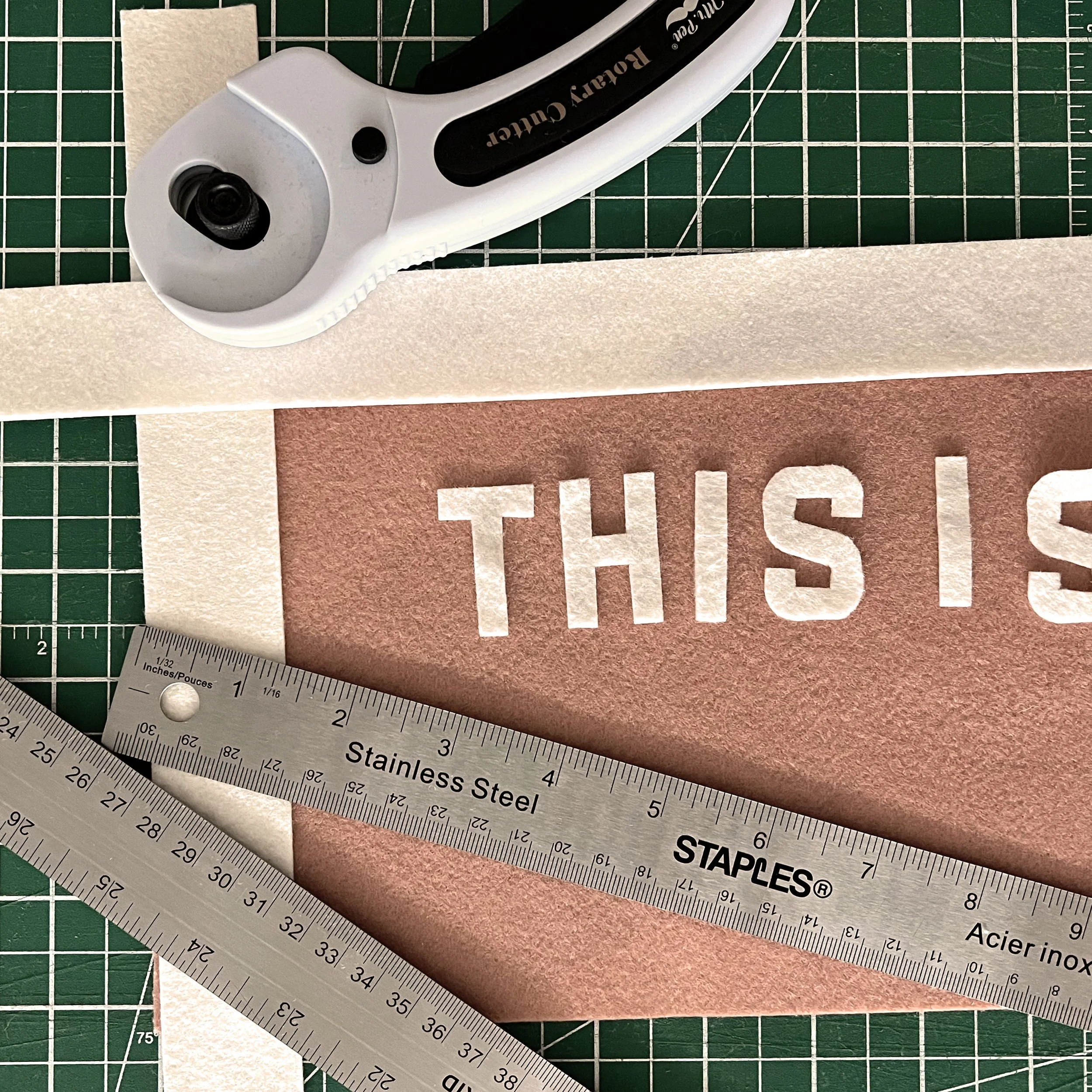 Close-up of a rotary cutter tool placed on a green cutting mat with grid lines, beside a stainless steel ruler and cut-out letters spelling 'TILE' and 'STAIRS' on colored paper.