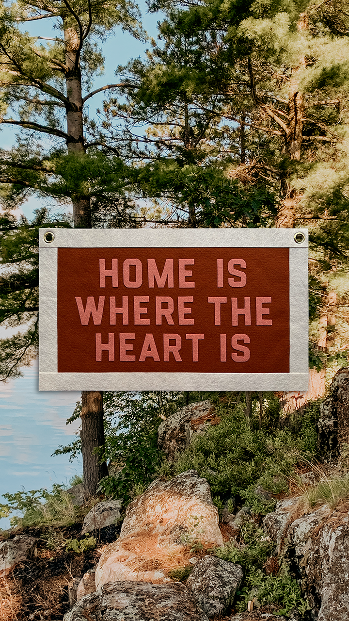 A sign that reads "Home is where the heart is" is attached to a tree among rocks and greenery, with tall pine trees and a blue sky in the background.