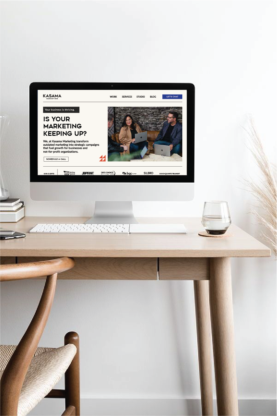 A computer monitor displaying a website for Kasama Marketing on a minimalist wooden desk with a glass of water, a keyboard, and a mouse, in a bright white room.