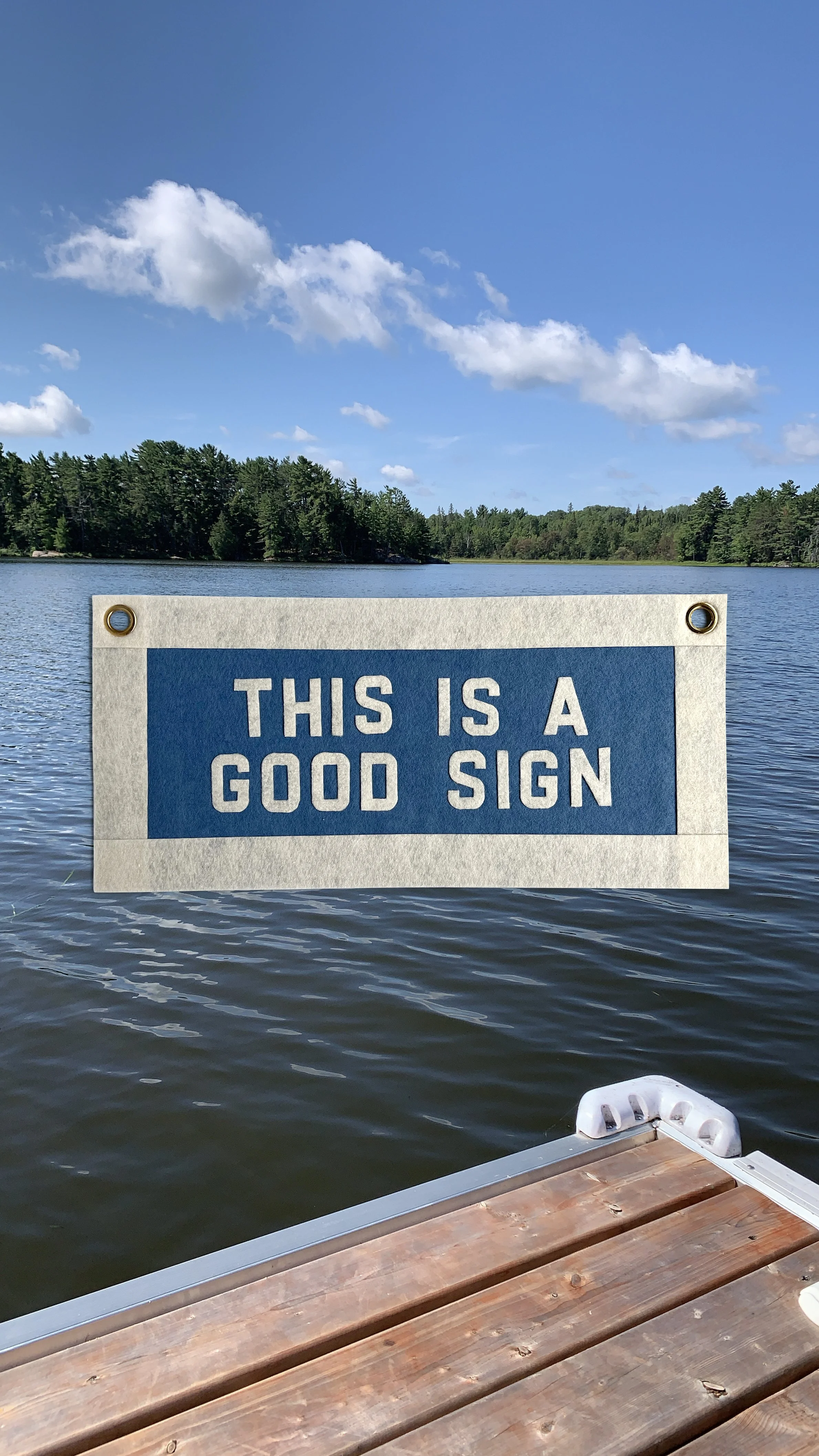 A sign on a lake reading 'This is a good sign' with a lakeside view, a clear blue sky, and trees in the background. Part of a dock and boat are visible in the foreground.