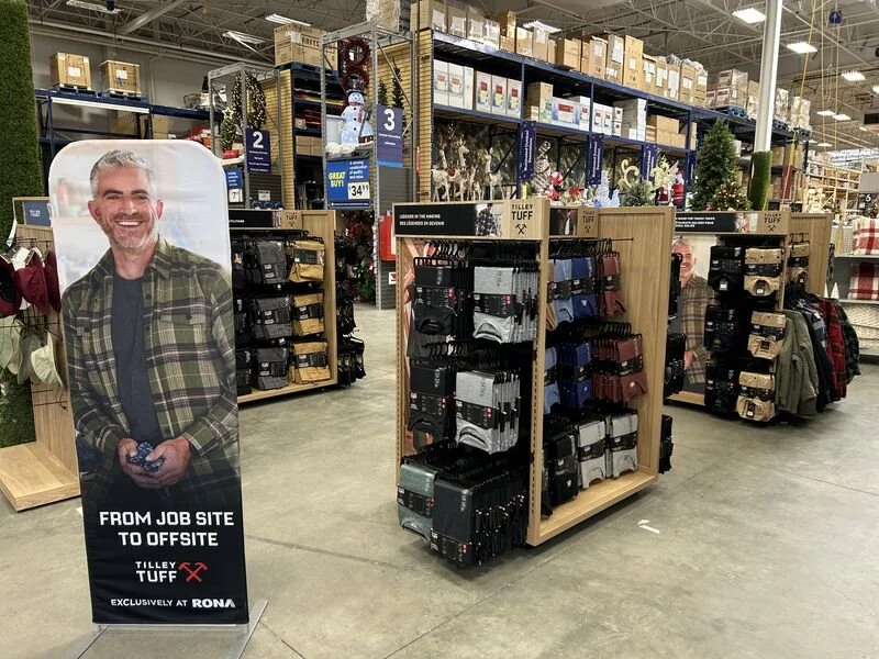 A retail store aisle with clothing and accessories on display. A standee features a smiling man with gray hair and a plaid shirt, with the text "From job site to offsite" and the Tilley Tuff logo, indicating exclusive availability at RONA. The store has shelves stocked with boxes, holiday decorations, and various apparel.
