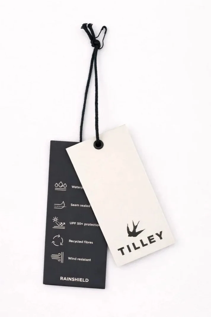 Clothing tags attached to fabric with string, featuring brand Tilley and Rainshield details like waterproof, seam sealed, UPF 50+ protection, recycled fibers, and wind resistant.
