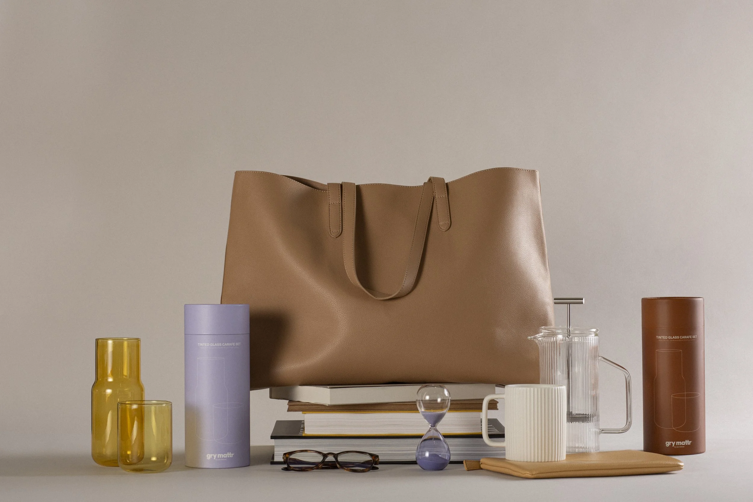 A beige leather tote bag is centered on a table, surrounded by various colorful glassware and containers, including yellow, purple, clear, and orange glasses, as well as stacked books and a pair of eyeglasses.