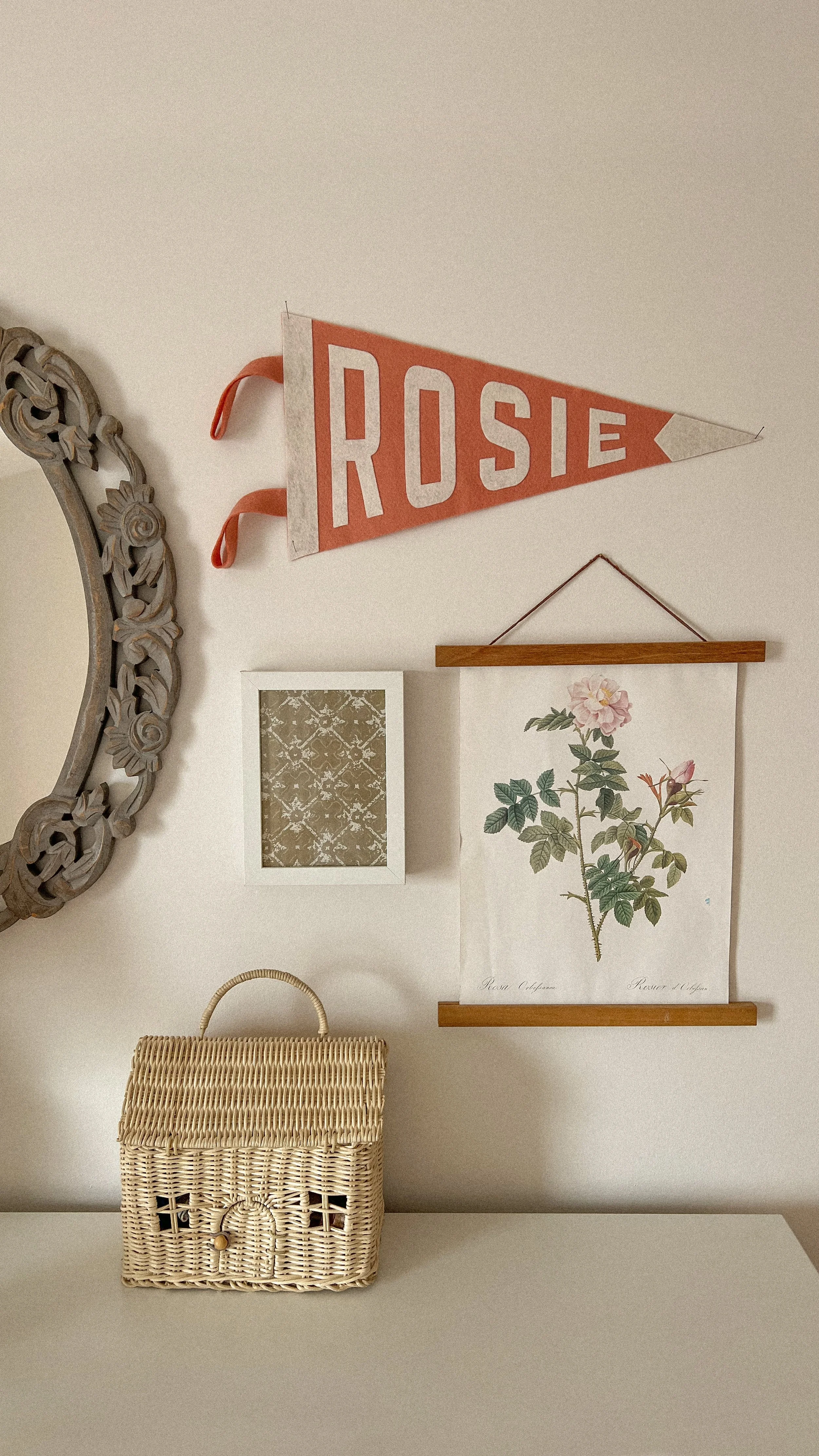 Decorative wall with a floral print, a side mirror with an ornate frame, a small frame with a patterned design, a hanging botanical print, a woven basket on a white surface, and a pennant banner with the name "Rosie".