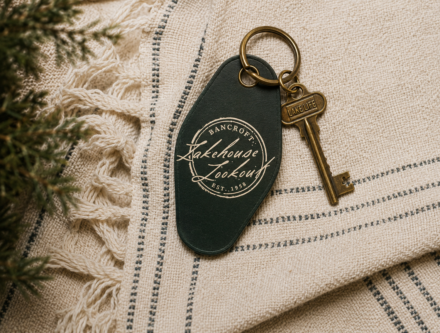 A set of keys with a black leather keychain that reads "Lakehouse Lockout," placed on a beige and blue striped woven fabric, with part of a green pine branch visible on the left.