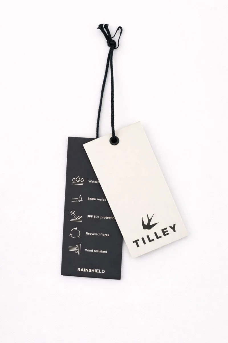 Two tags hanging from black string, one white with a bird logo and Tilley written underneath, the other black with sustainability features and Rainshield branding.
