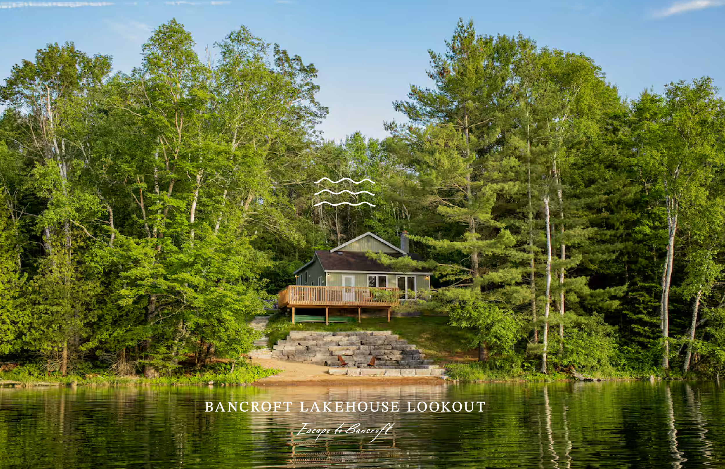 A lakefront scene with a house and deck surrounded by tall green trees, featuring stone steps leading down to the water and the text "Bancroft Lakehouse Lookout" at the bottom.