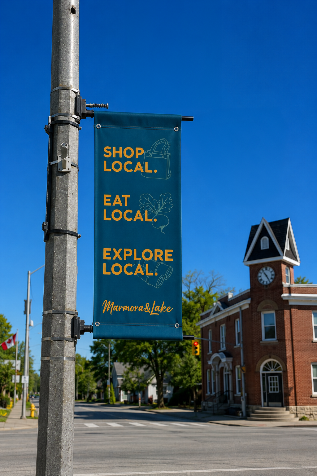 A vertical blue banner on a street pole reads 'Shop Local. Eat Local. Explore Local.' with drawings of a shopping bag, leafy vegetable, and roll of paper, and the text 'Marmora & Lake' at the bottom. In the background, there are trees, a building with a clock tower, and a traffic light against a clear blue sky.