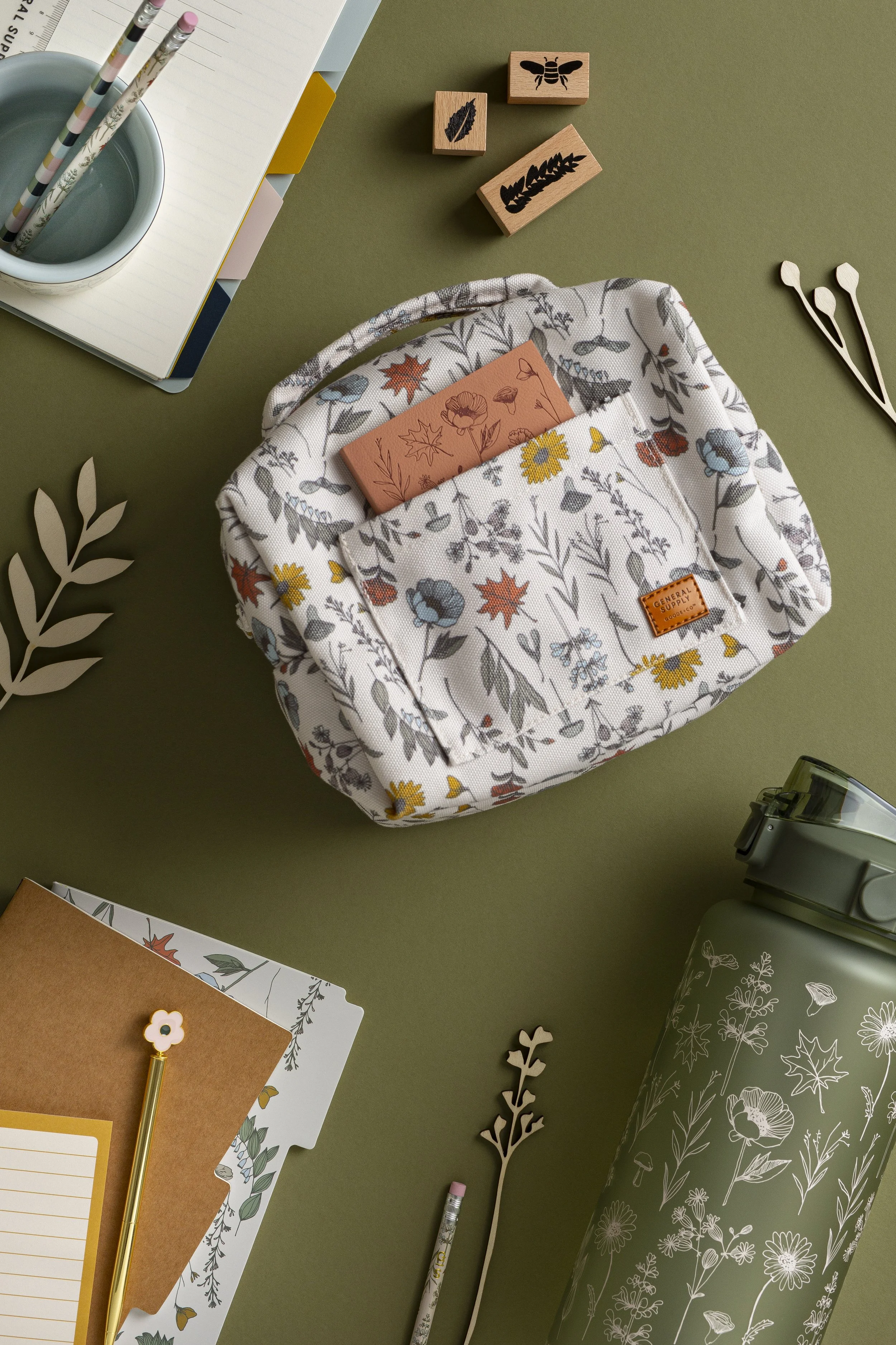 A flat lay of stationery and office supplies on a green surface. Items include notebooks, pencils, paper clips, decorative stamps, a floral-patterned tote bag, a water bottle, and loose paper with botanical designs.