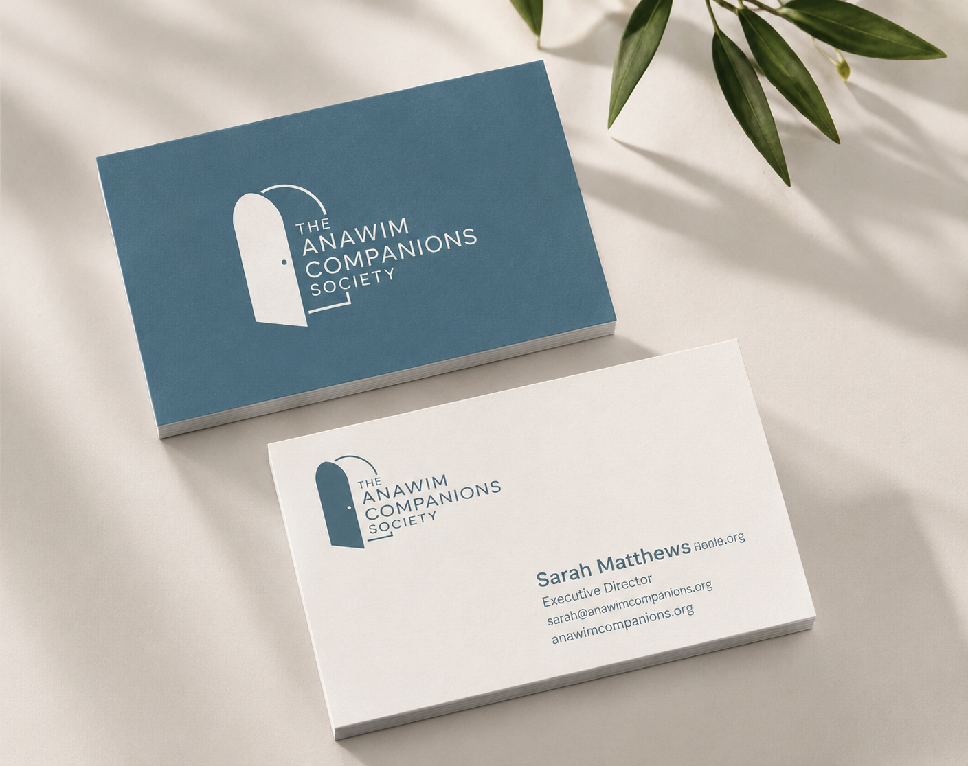 Business cards for The Anawim Companions Society, with the logo featuring a stylized door and the organization's name, one card showing the organization’s logo and the other displaying a person's contact information including name, email, and title, placed on a light surface with a green plant nearby.