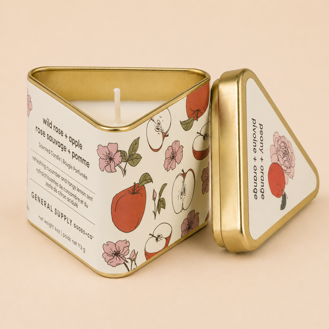 A decorative candle tin with a lid, featuring floral and fruit illustrations, labeled with scent descriptions 'wild rose + apple' and 'peony + orange pivoine + orange', and containing a white candle wick inside.