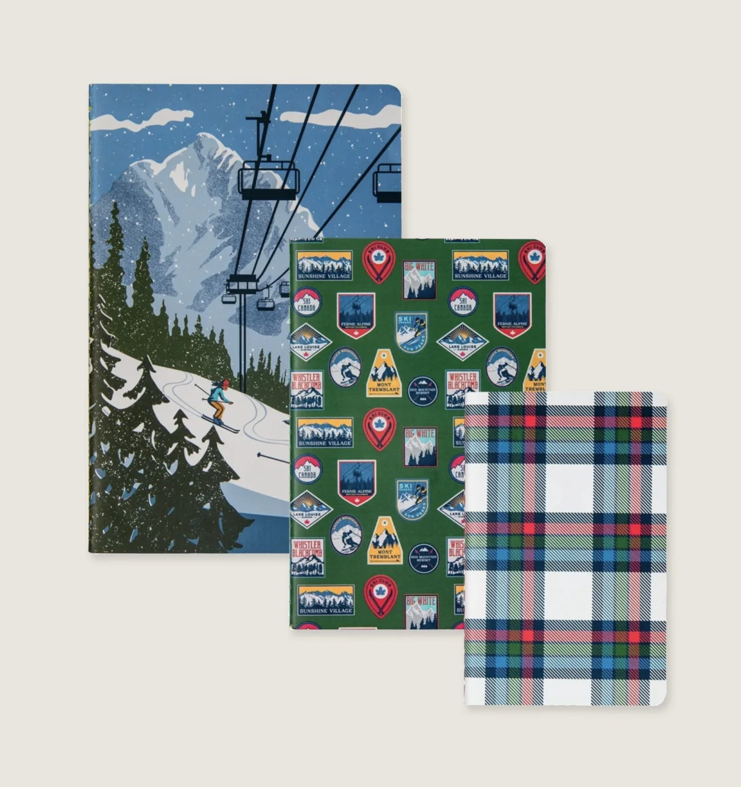 Three notebooks with ski resort themes, one featuring a snowy mountain scene, one with colorful patches of various ski resort logos, and one with a blue, red, green, and white plaid pattern.