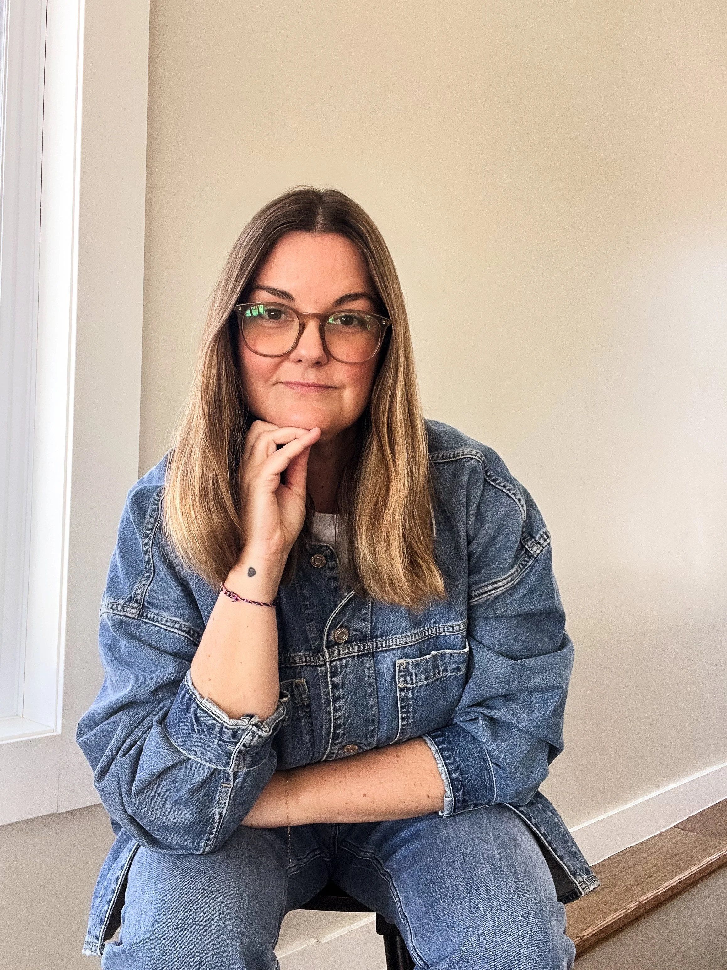 A woman with long brown hair and glasses sitting on a staircase, wearing a denim jacket and jeans, resting her chin on her hand, in front of a plain beige wall.