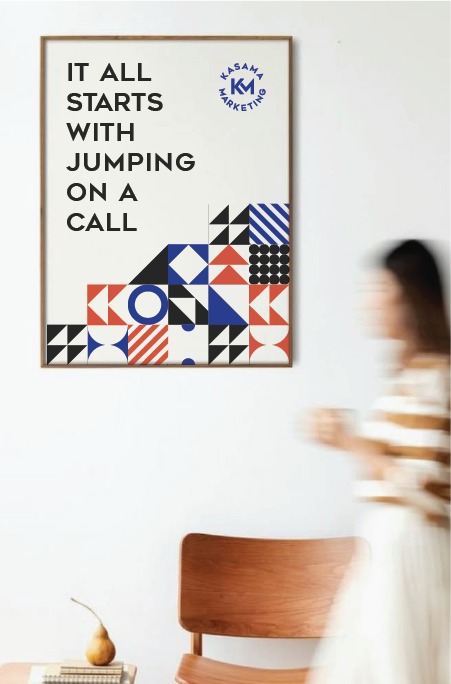 Framed poster on a white wall with the quote 'It all starts with jumping on a call' and geometric patterns in black, white, red, and blue. Blurred woman walking past in a striped top. Wooden chair and small decorative plant on a table below the poster.