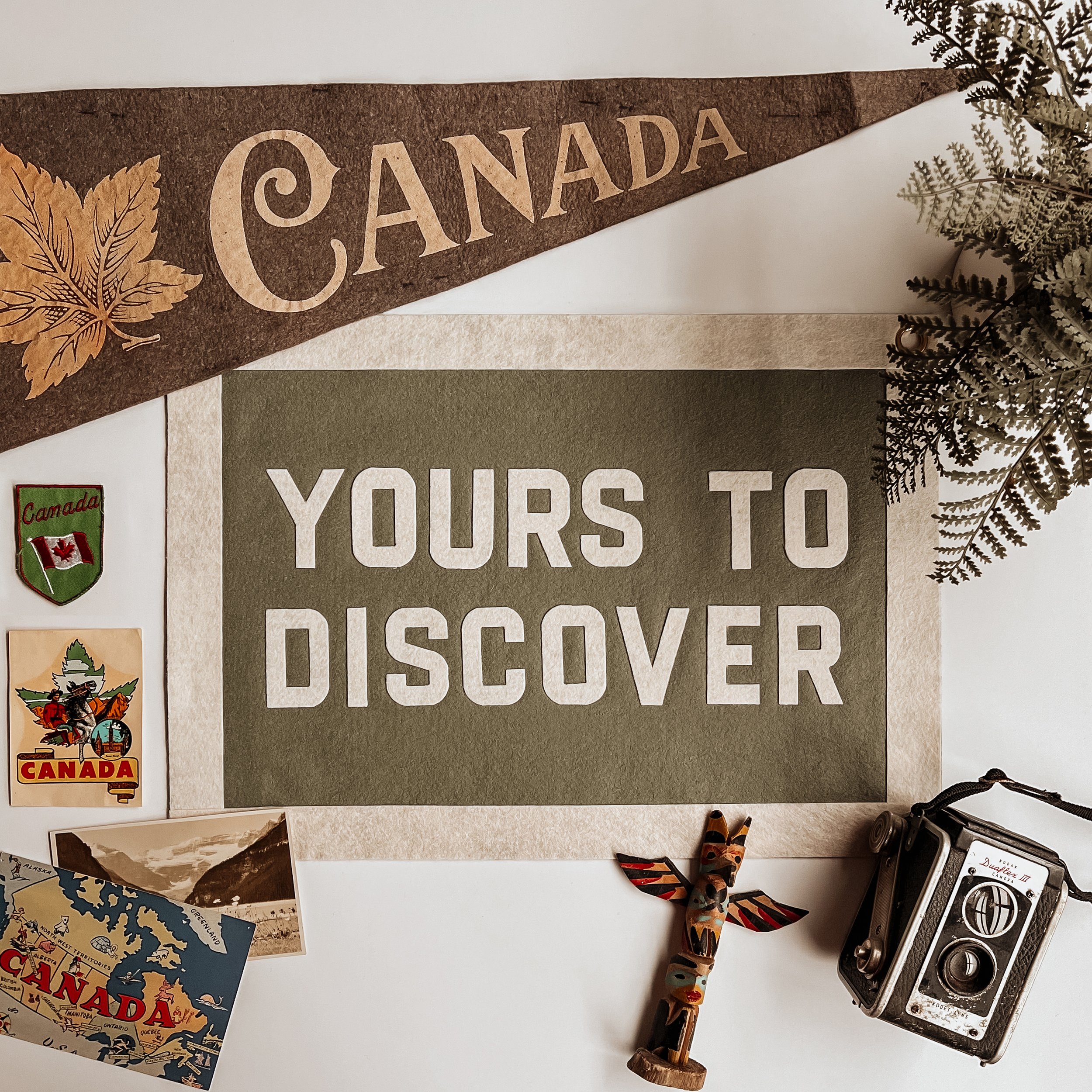 Decorative sign with the word 'Canada' and a leaf graphic, part of a collection of travel souvenirs including maps and a vintage camera, with a pair of wooden earrings or figurines on a white surface.