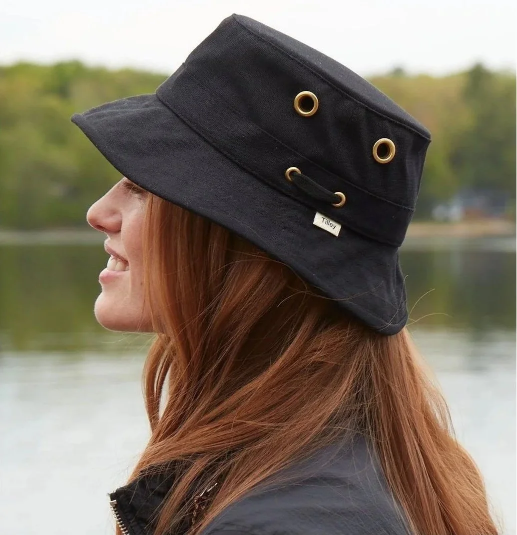 Side profile of a woman with long red hair wearing a black wide-brimmed hat with gold grommets and a black jacket, outdoors near a body of water with a blurred green and brown background.