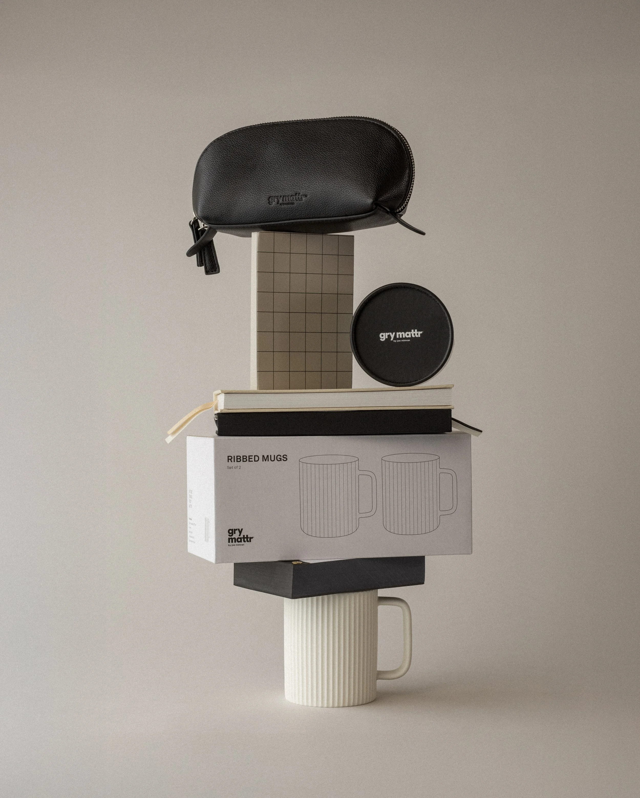 Multiple objects on a beige background, including a black leather zippered pouch with the word "gry matt" on it, a black round container with white text, a white box labeled "RIBBED MUGS," a tall rectangular object with a grid pattern, and various books and bookshelves.