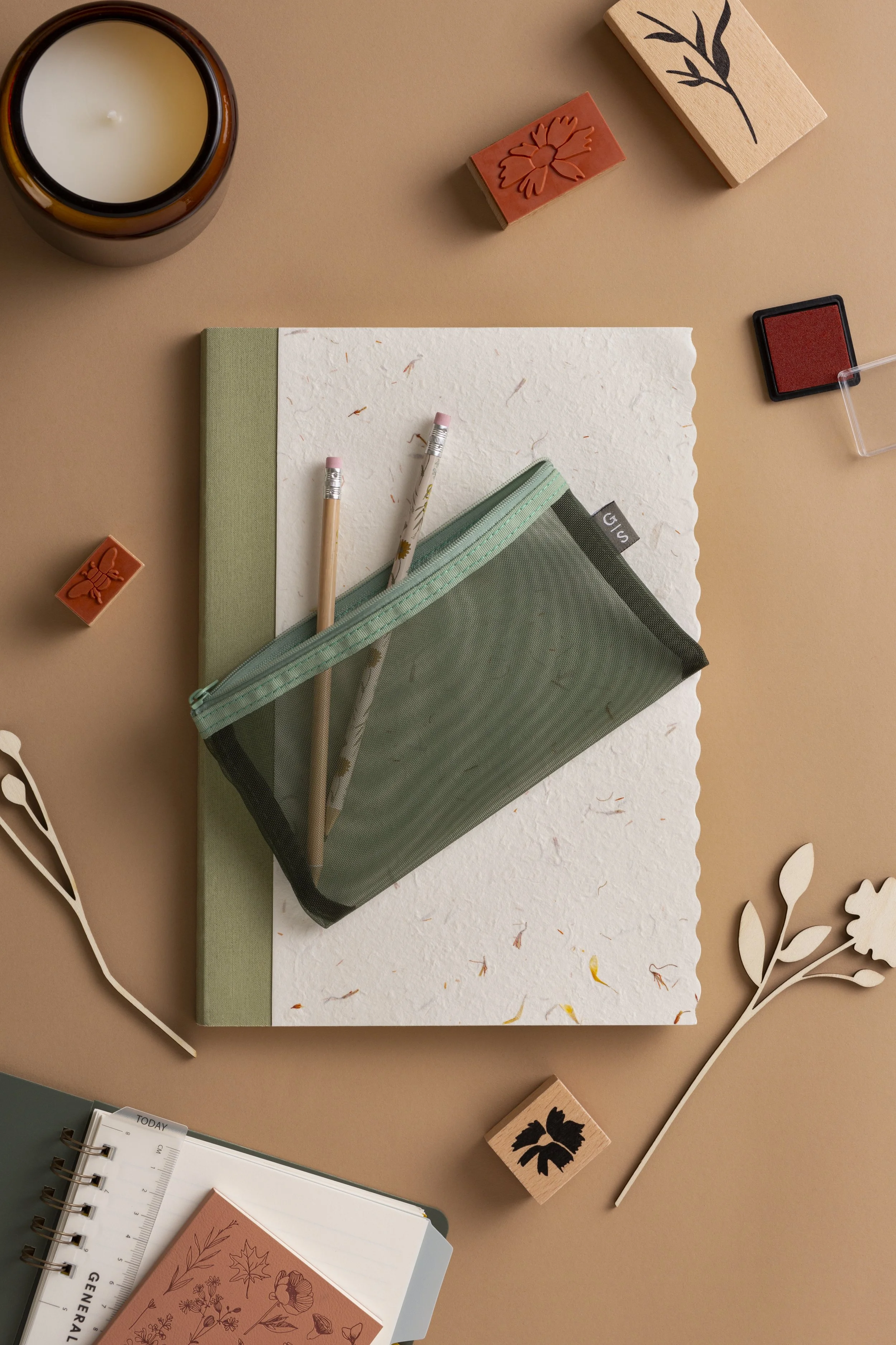 A flat lay of stationery items, including a green and white textured notebook with a zippered pouch, two pencils with floral and daisy designs, a red and pink ink pad, wooden stamps with leaf and flower designs, a white paper doily, and a candle in a brown glass holder on a peach-colored background.