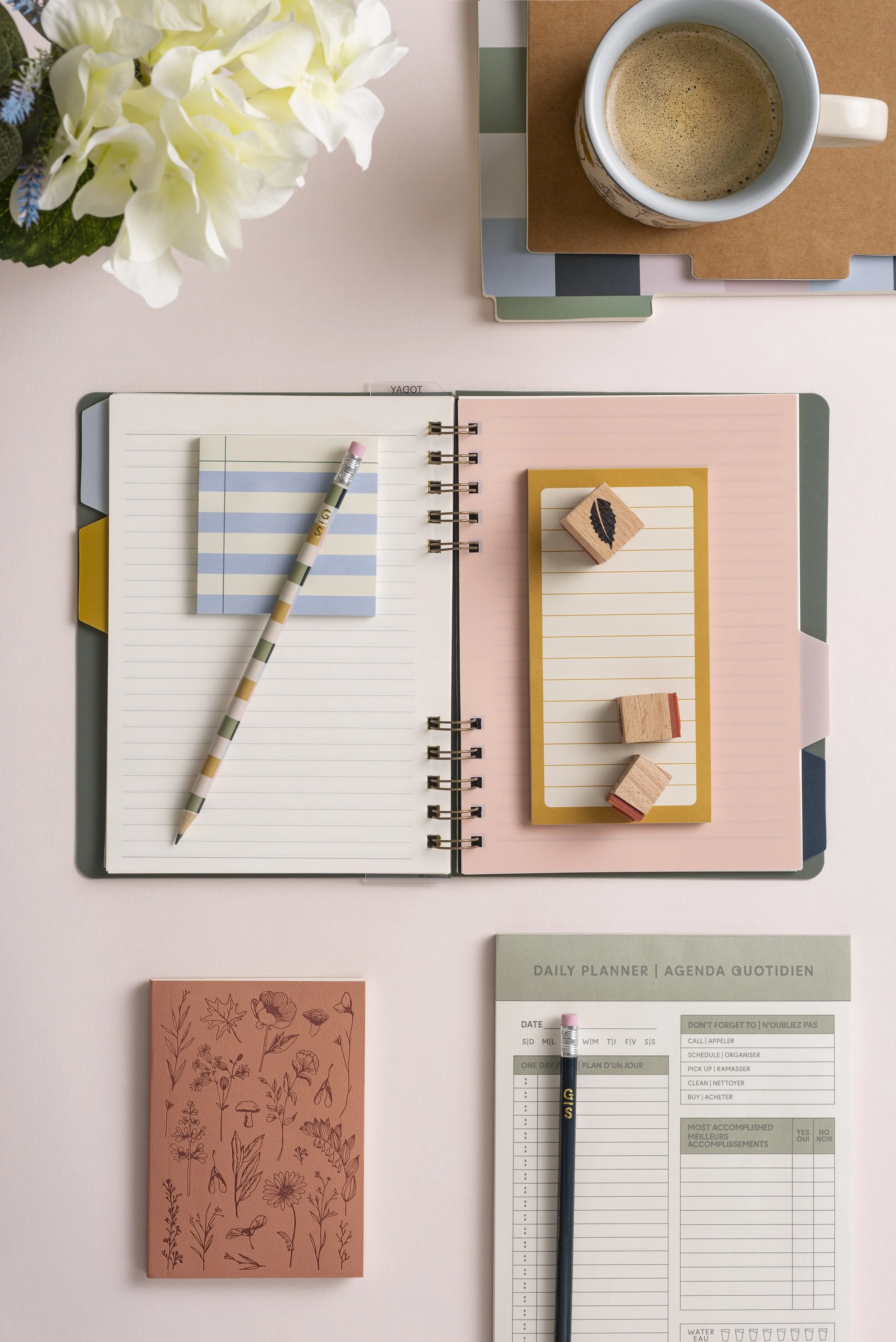 Flat lay of a pink desk with a white open planner, lined paper, sticky notes, wooden stamps, a pen, a mug of coffee, a floral notepad, and a notebook with floral illustrations, flowers, and a daily planner sheet.