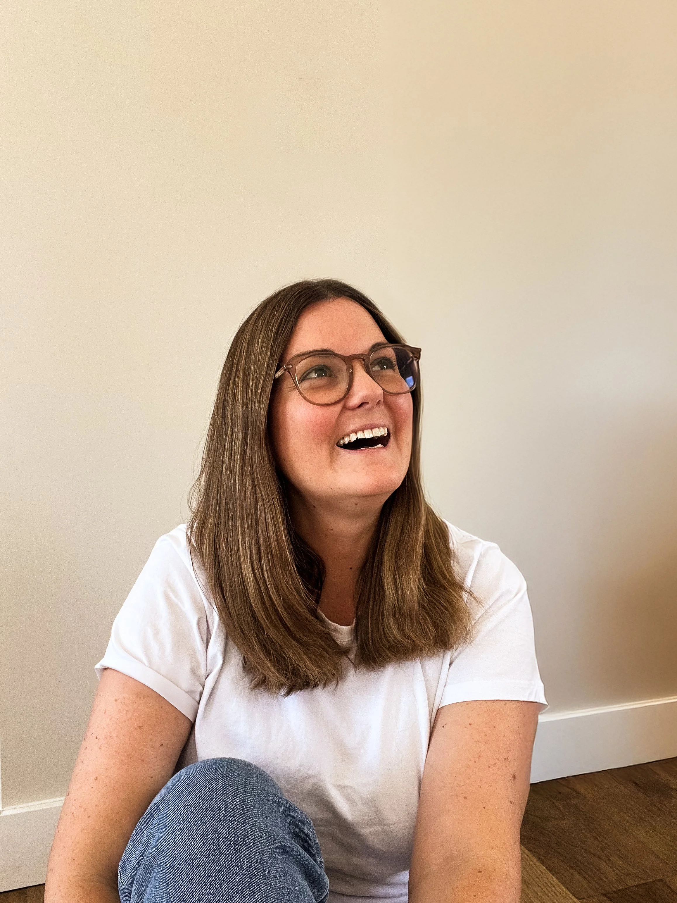 A woman with long brown hair, glasses, and freckles, sitting on the floor, smiling, wearing a white t-shirt and blue jeans, against a plain beige wall.