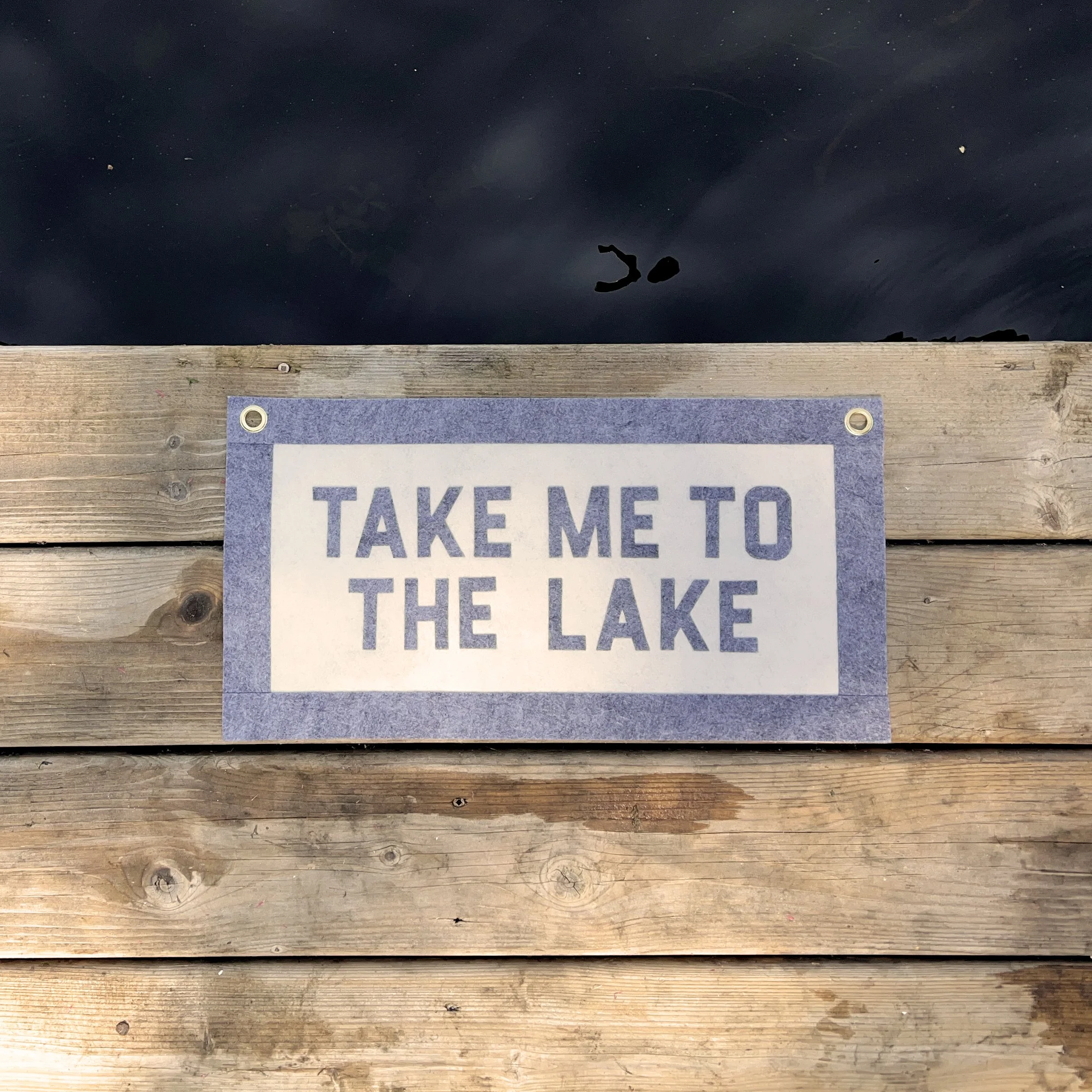 A wooden surface with a partially visible sign that reads 'TAKE ME TO THE LAKE' and a dark background with a reflection of a person or object.