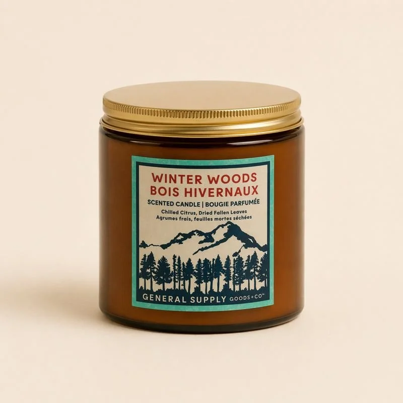 A brown glass candle container with a gold lid, labeled Winter Woods with a mountain and pine tree illustration.