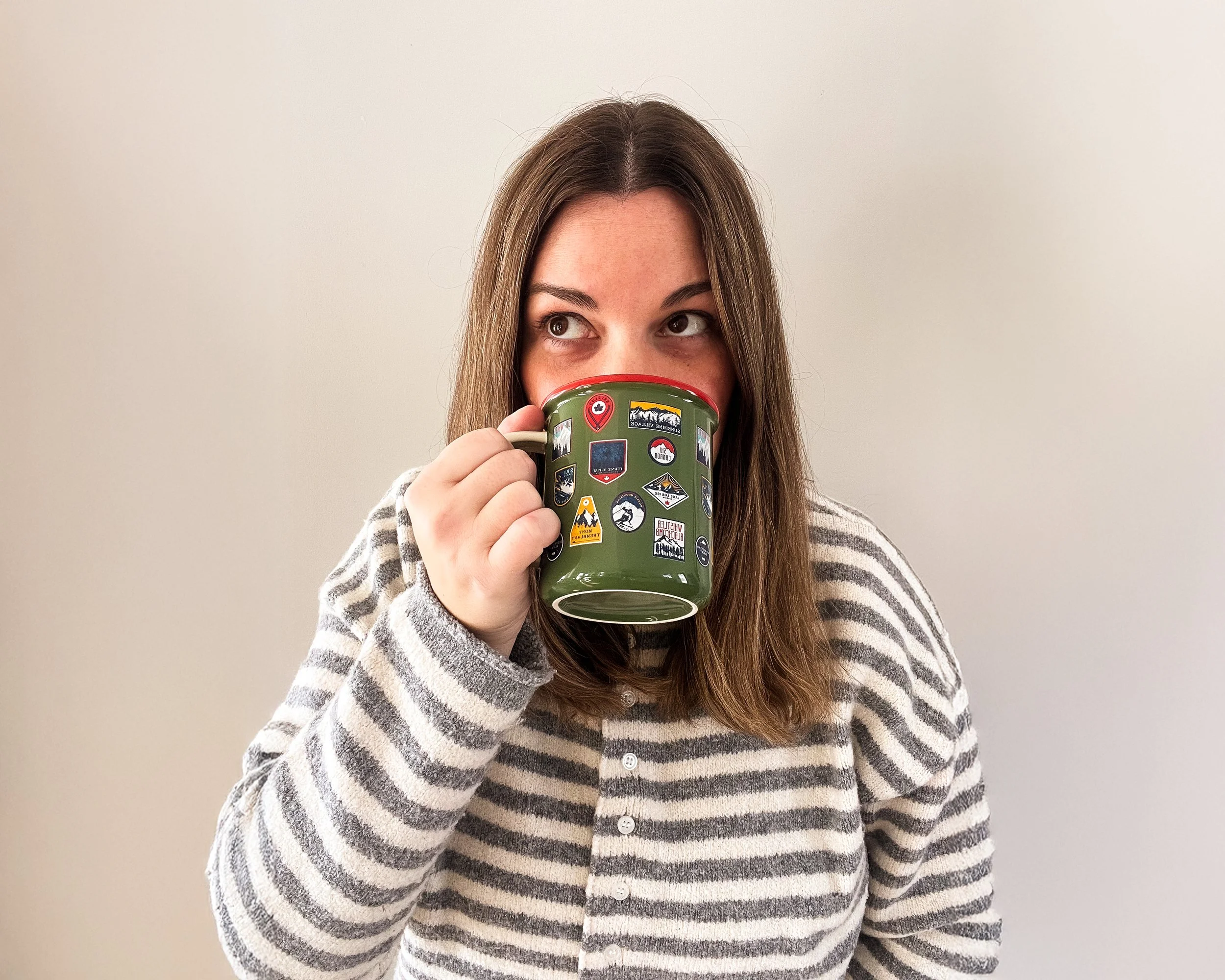A woman with long brown hair and light skin holding a green mug with outdoor and mountain stickers, wearing a gray and white striped shirt, against a plain white background.
