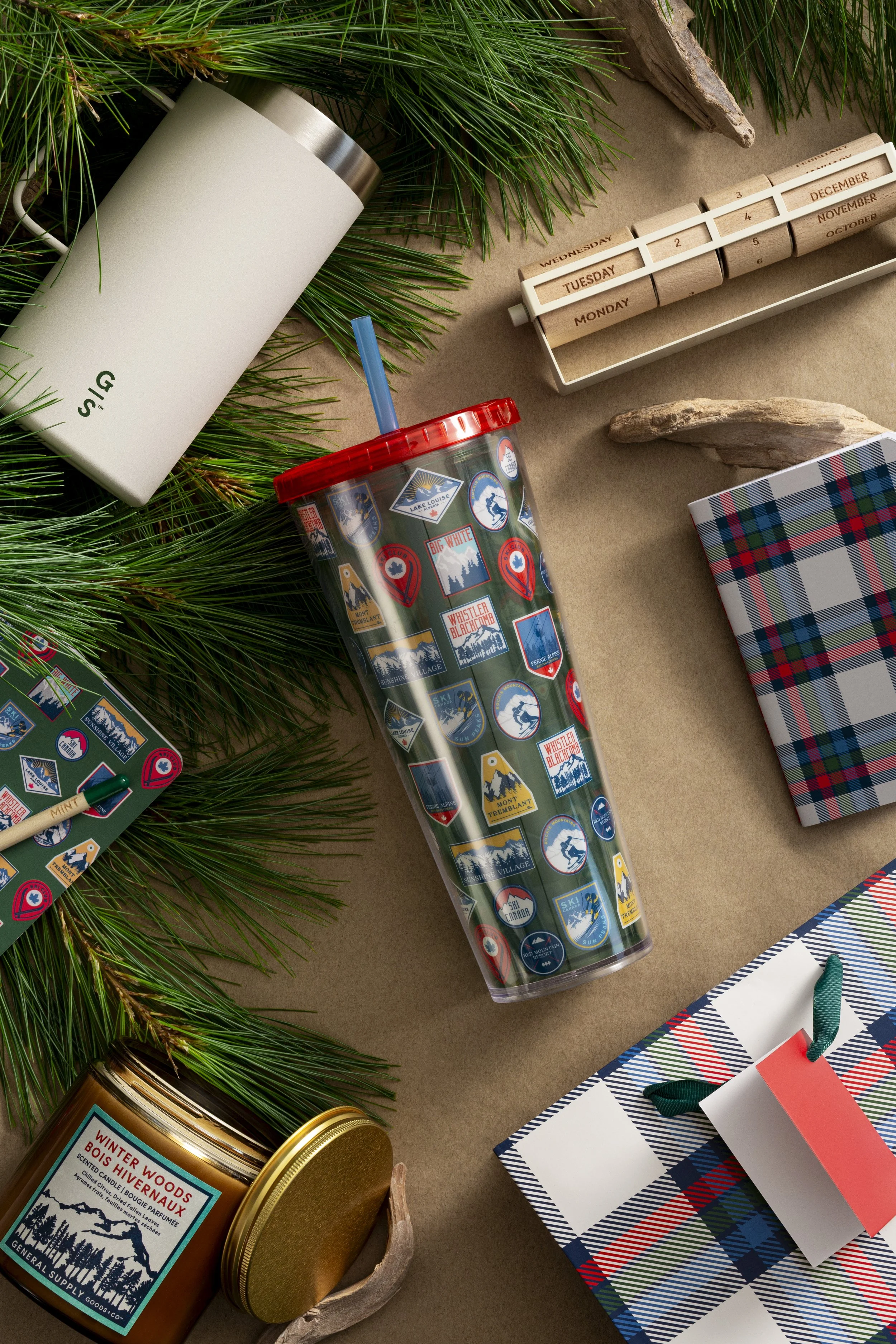 Assorted holiday and winter-themed items including a white insulated tumbler, a colorful travel mug, a pine branch, wooden desk calendar displaying days of the week and months, a wrapped gift box with plaid pattern, a gift bag with ribbon handles, a scented candle, and a piece of driftwood, all set on a brown background.