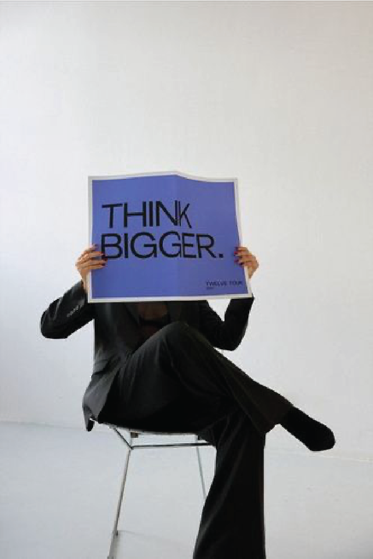 Person in black clothing sitting on a chair, holding a blue sign that says "THINK BIGGER." in front of their face, against a plain white background.