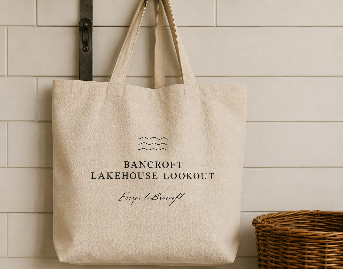 A beige tote bag hanging on a hook against a white tiled wall, with the text "Bancroft Lakehouse Lookout" and "Escape to Bancroft" printed on it. A wicker basket is partially visible at the bottom right.
