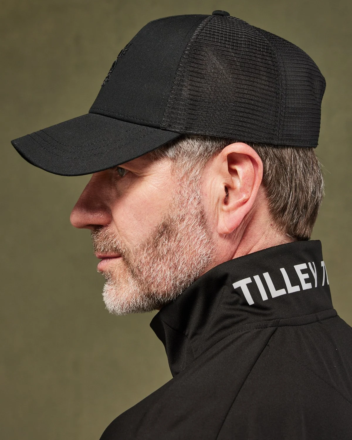 Profile view of an older man wearing a black baseball cap and black jacket with white text on the collar.
