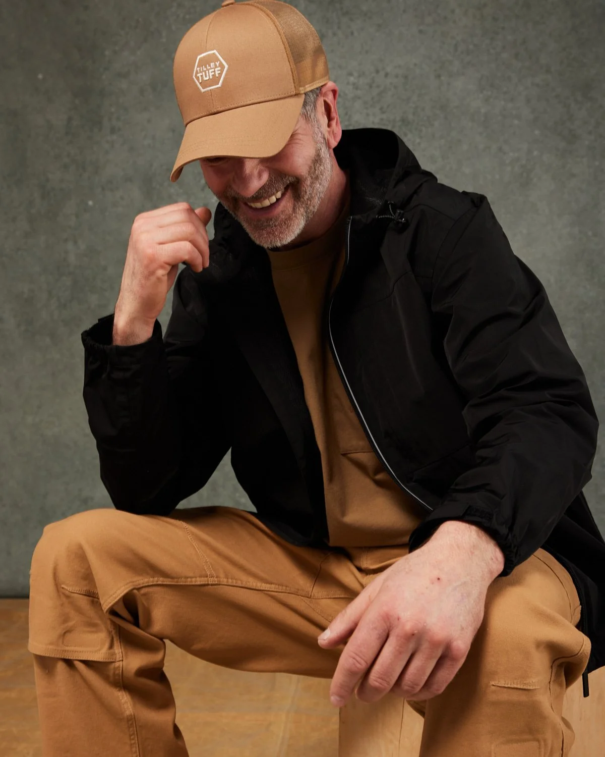 A man laughing, wearing a tan cap with a "Tilley Tuff" logo, a black jacket, and tan pants, sitting against a gray wall.