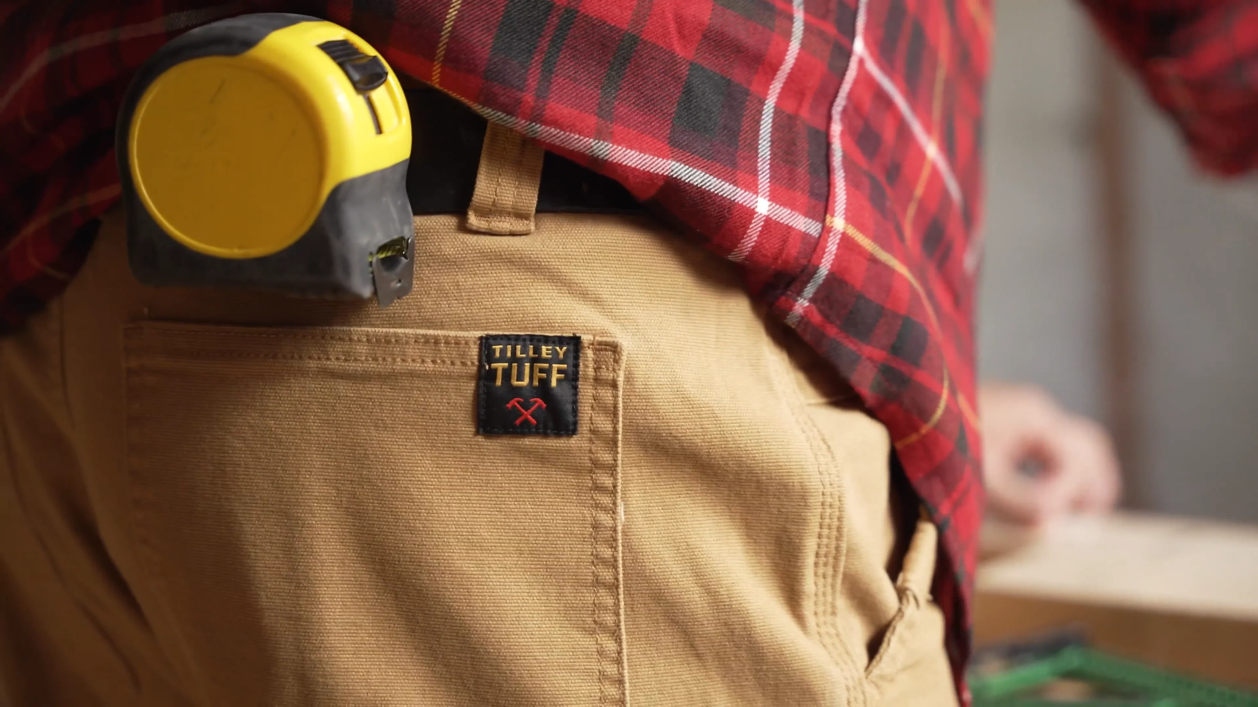 Close-up of a person wearing tan work pants with a 'Tilley Tuff' label, a red plaid shirt, and a yellow measuring tape hanging from the pocket.
